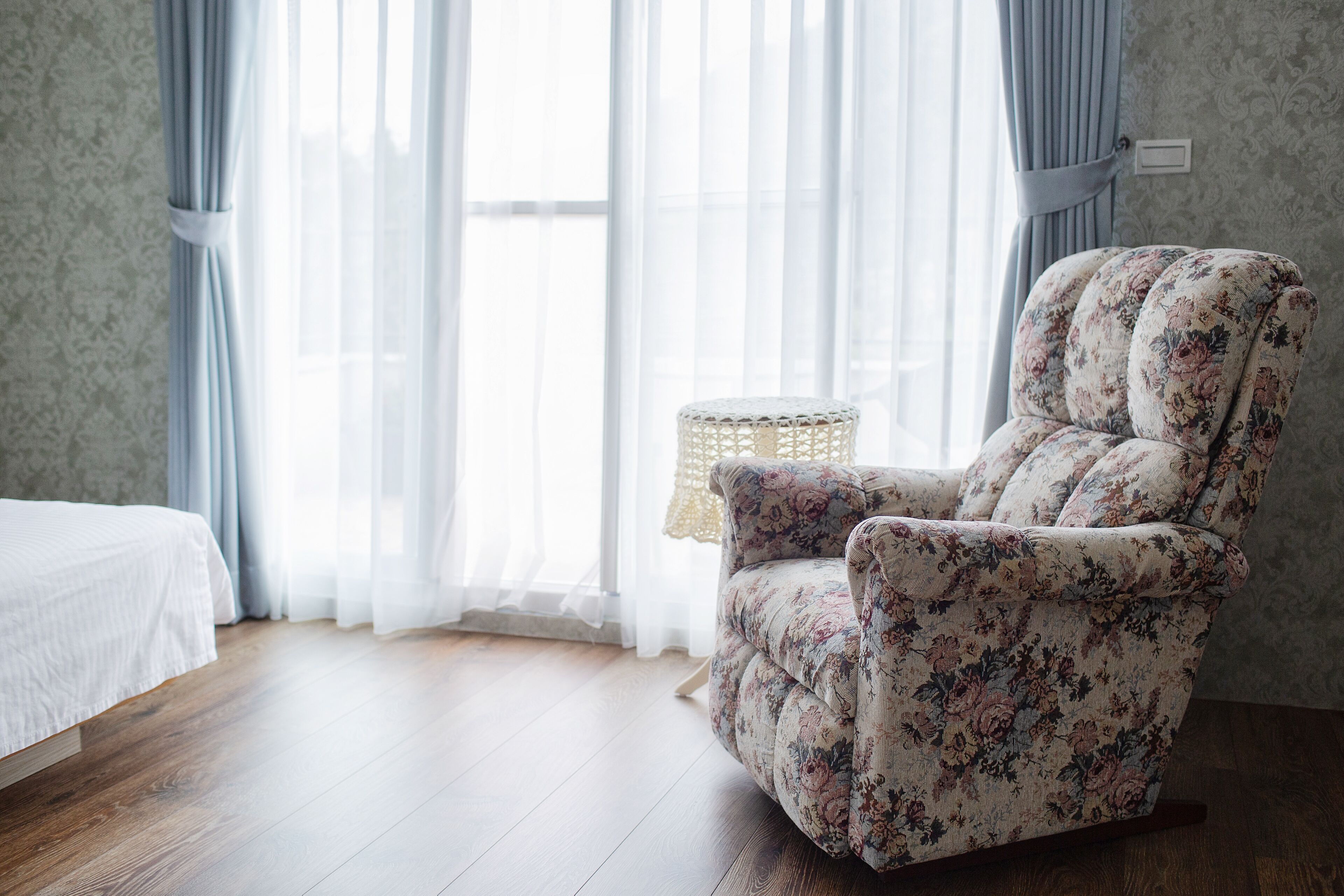 A room with a floral-patterned armchair, a bed with a white duvet, and a window with sheer curtains.