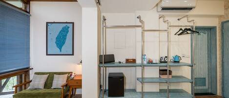 A room with a wooden floor, a green sofa, a desk with a computer, a small black refrigerator, and a blue hanging rack with clothes.
