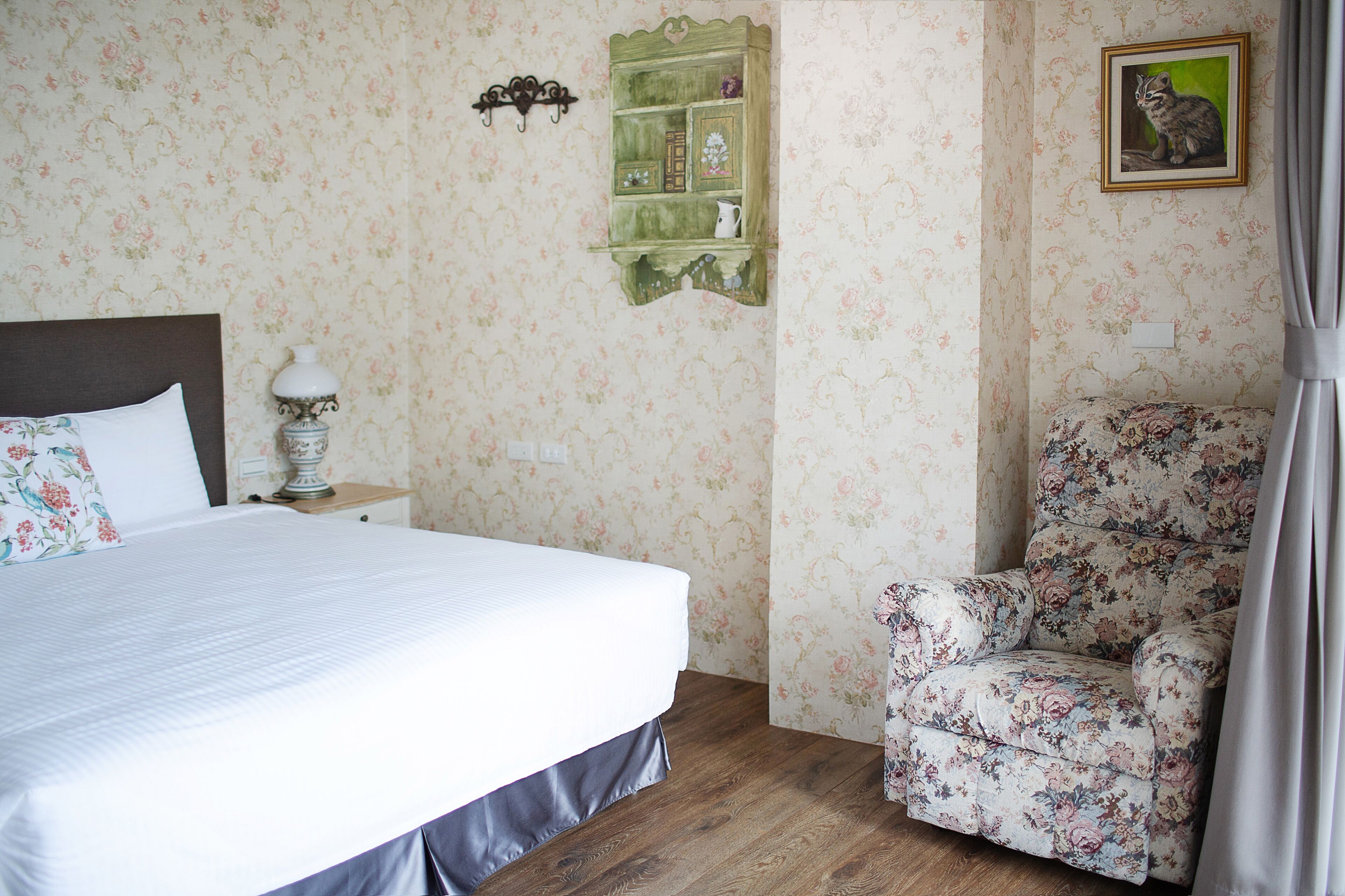 A neatly arranged bedroom with a bed, a nightstand, a floral armchair, and a green shelf unit.
