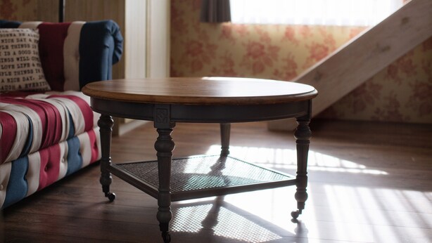 Una mesa redonda de madera con base de malla metálica en una habitación con papel tapiz estampado y un sofá a rayas.