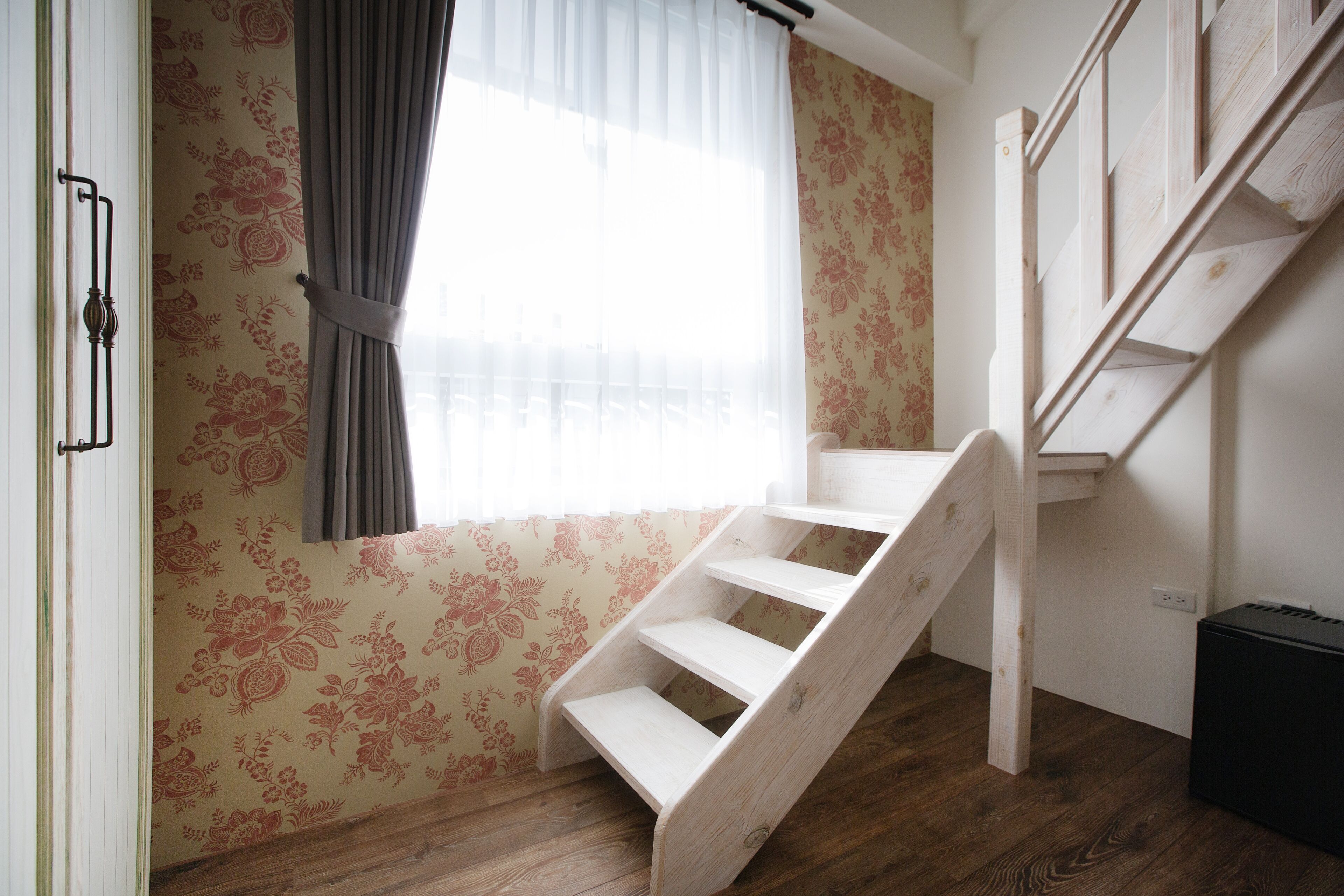 A room with a wooden staircase, floral wallpaper, and a window with curtains.