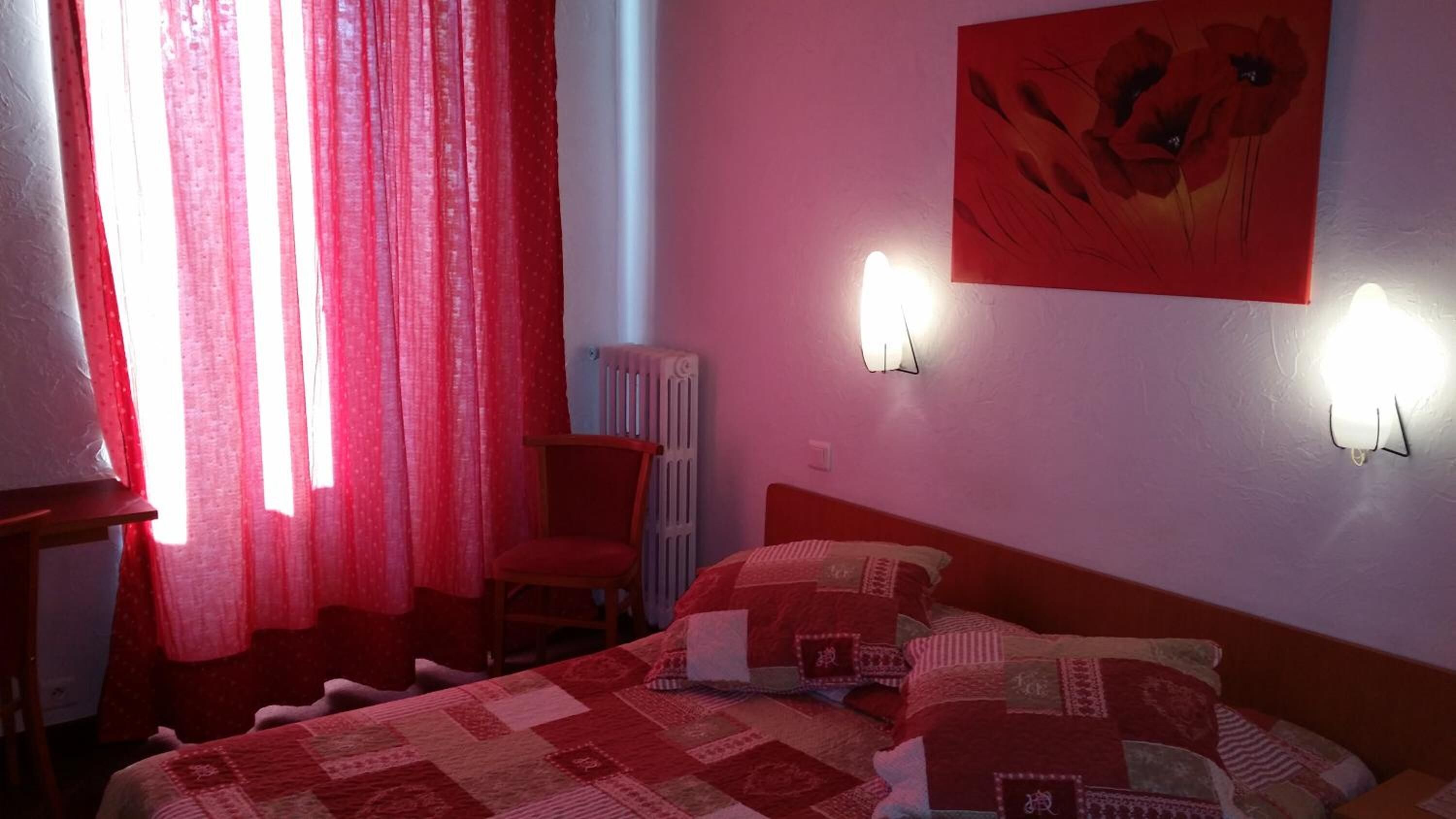 Un dormitorio con colcha de cuadros rojos y blancos, una silla, un radiador y una pared con dos apliques.