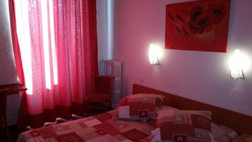 Un dormitorio con colcha de cuadros rojos y blancos, una silla, un radiador y una pared con dos apliques.