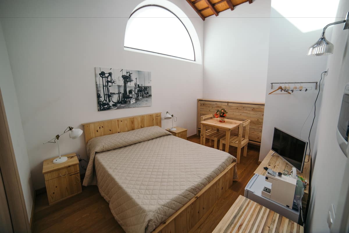 A bedroom with a bed, bedside table, lamp, and a desk with a chair. There is a window above the bed and a TV on a stand.