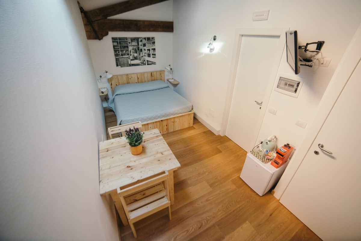 A compact room with a bed, a small table with a plant, a desk, and a door with a light switch and a small shelf.