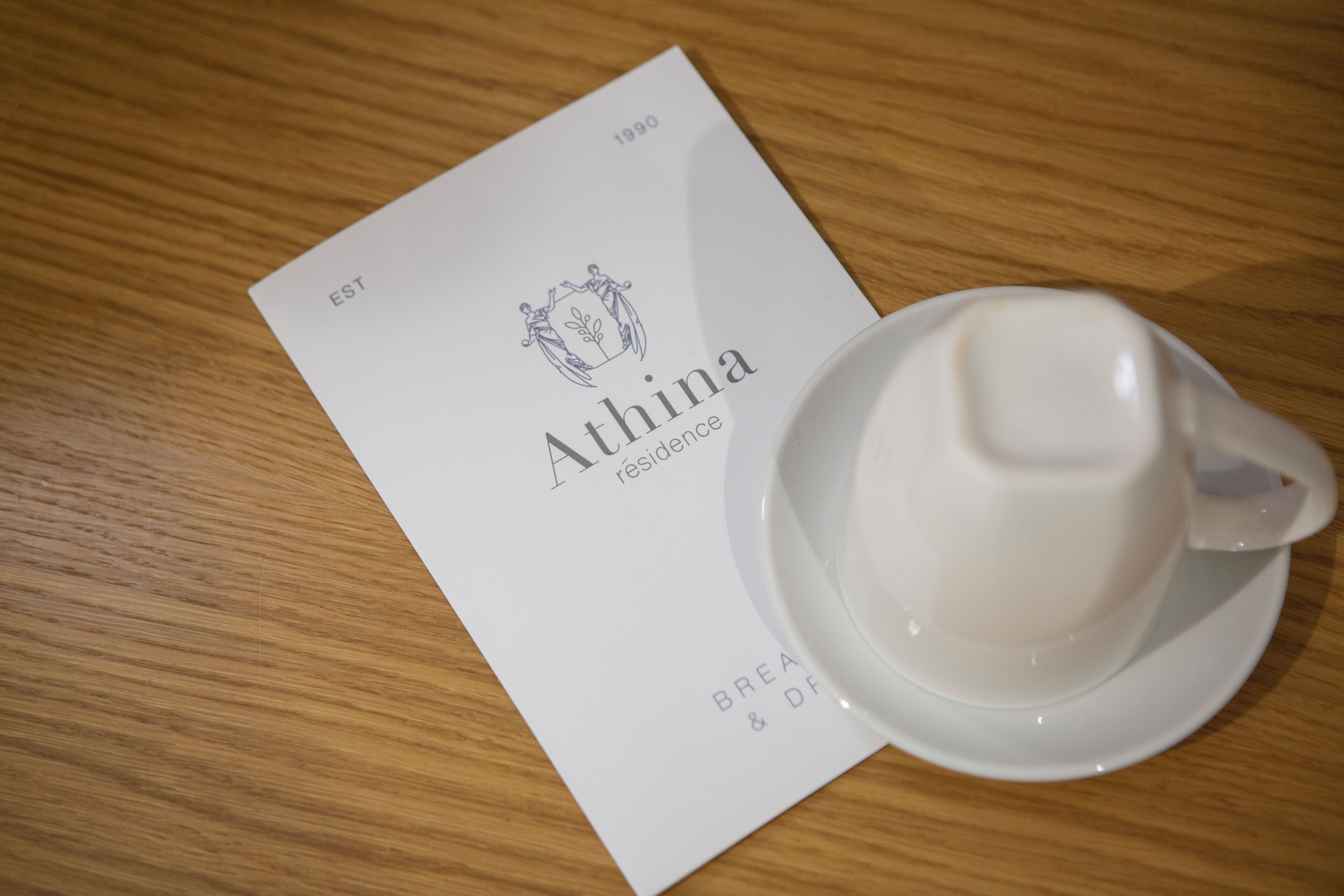 Une tasse blanche posée sur une soucoupe, à côté d’un menu sur lequel on peut lire les mots « Athina » et « 1890 ».