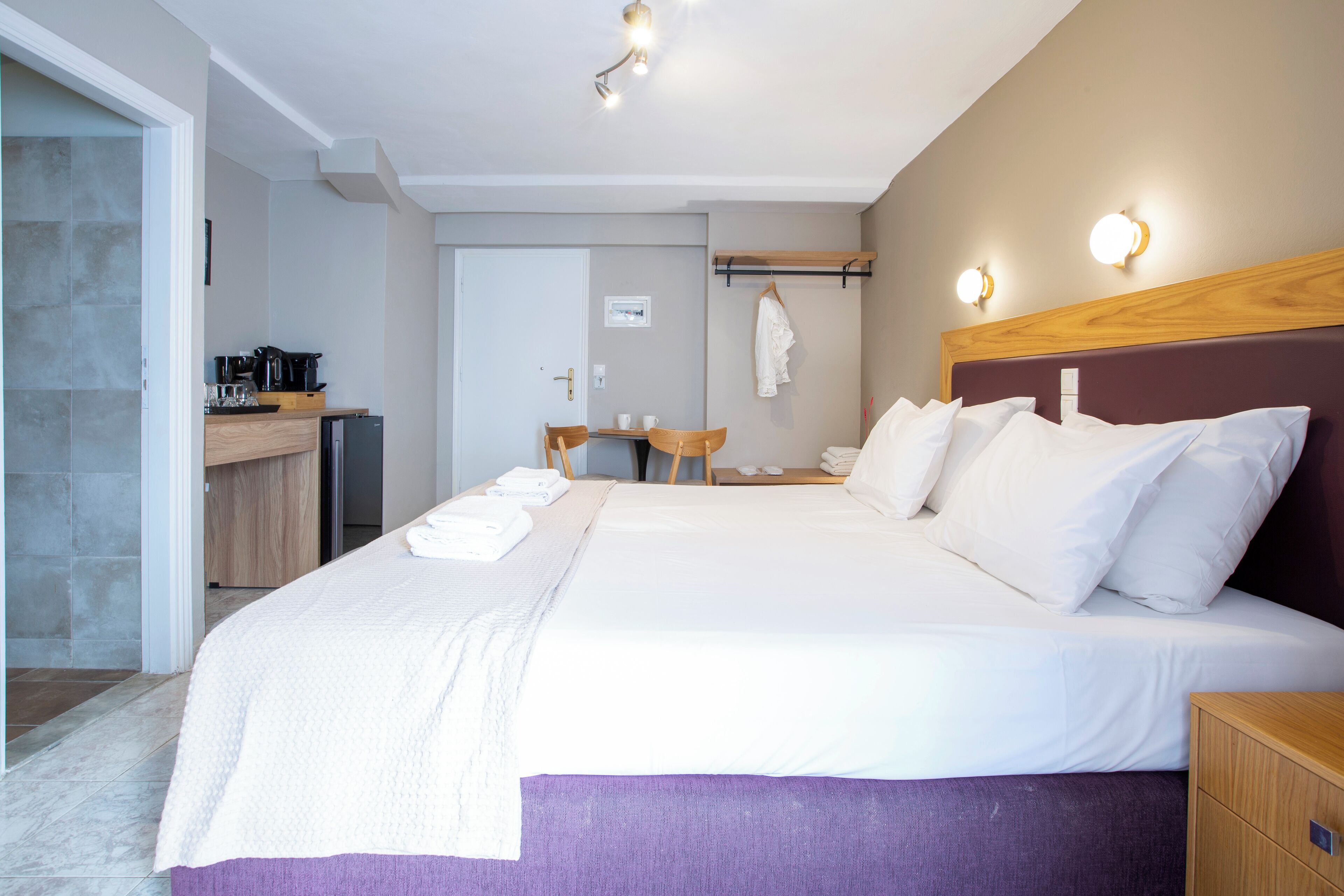A hotel room with a large bed, a wooden nightstand, a desk with a chair, and a small kitchenette.