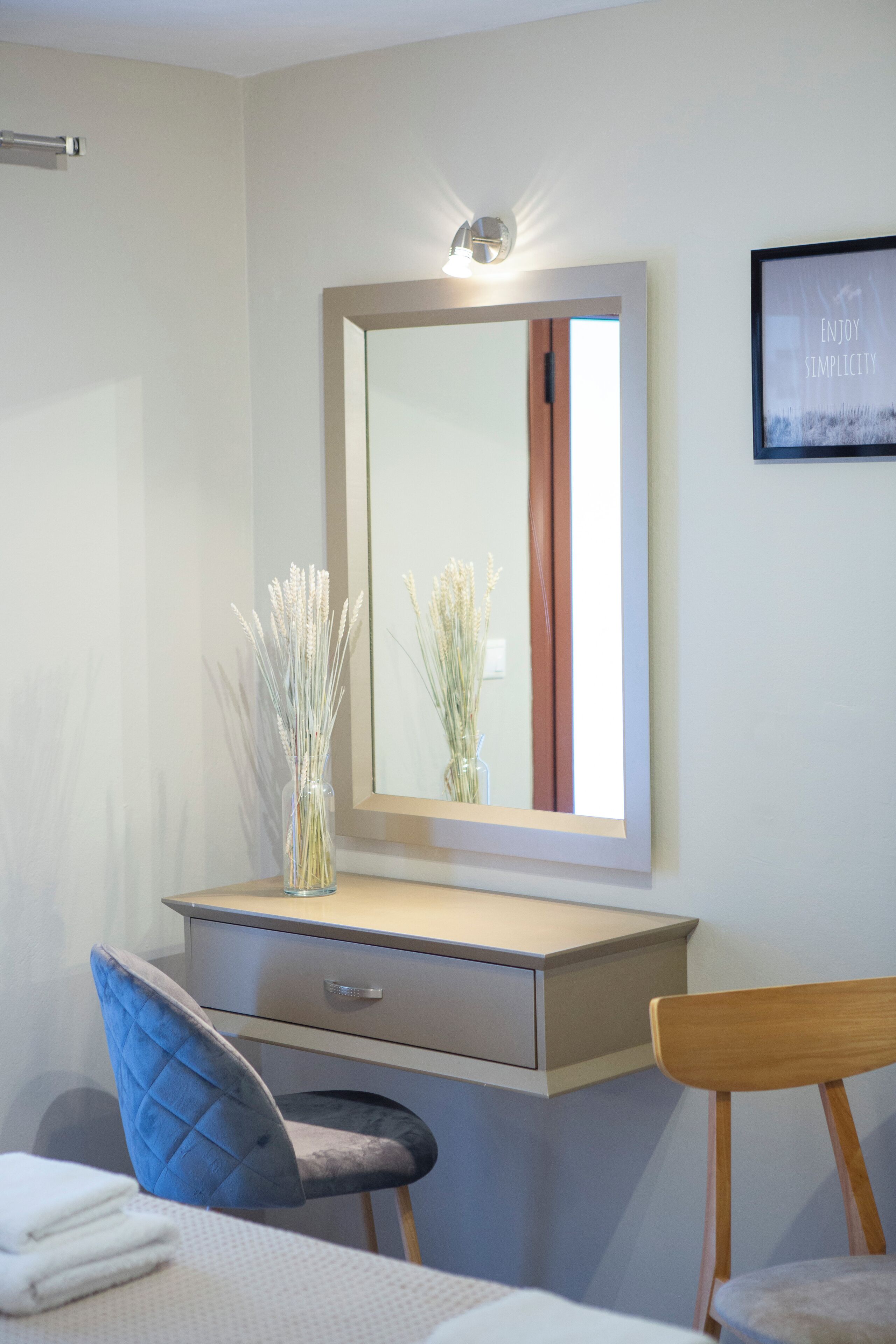 Habitación de hotel con tocador, espejo, florero con flores secas, una silla azul y una silla de madera.