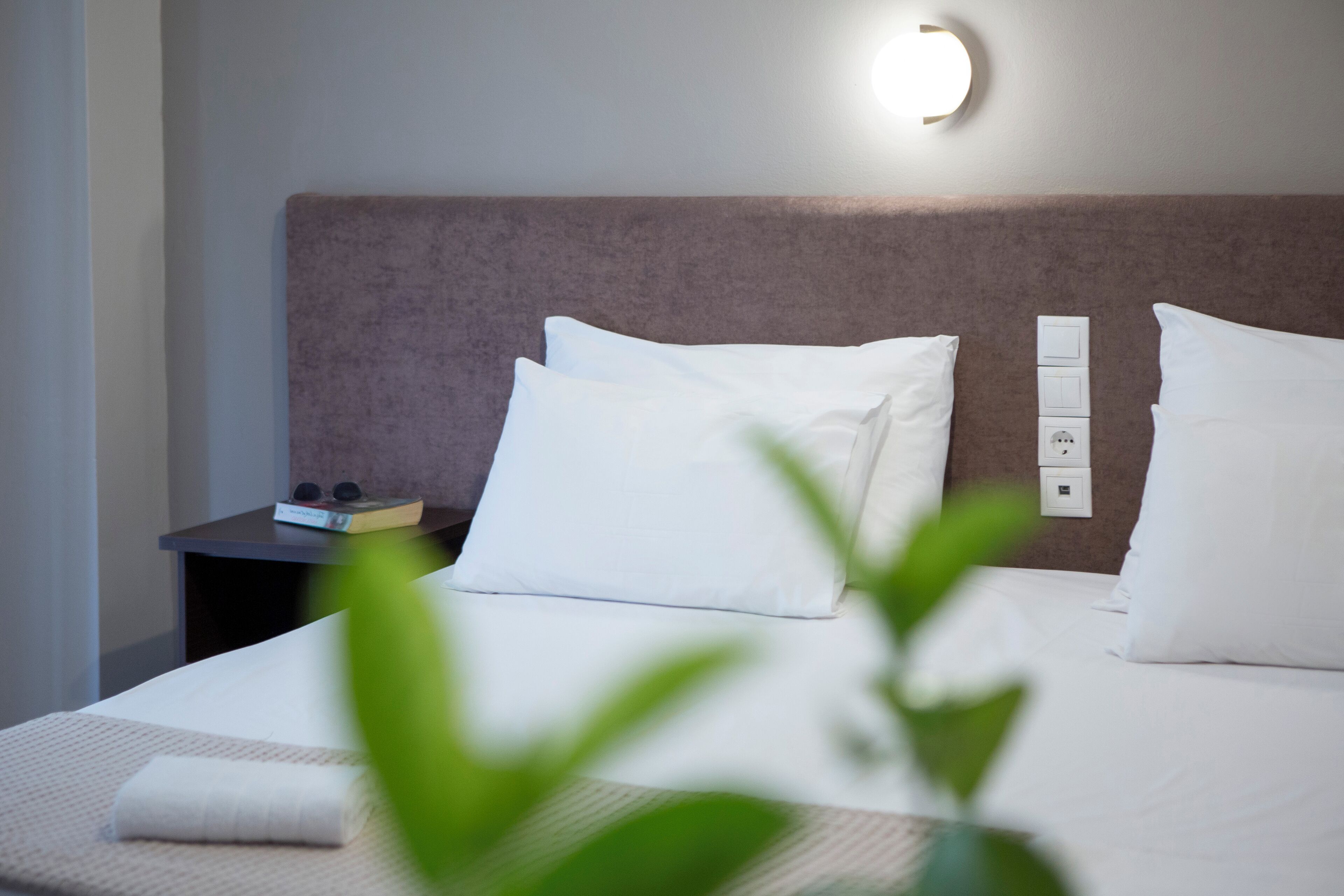 A neatly made bed with white linens and pillows, a bedside table with a book and a small electronic device, and a wall-mounted light fixture.