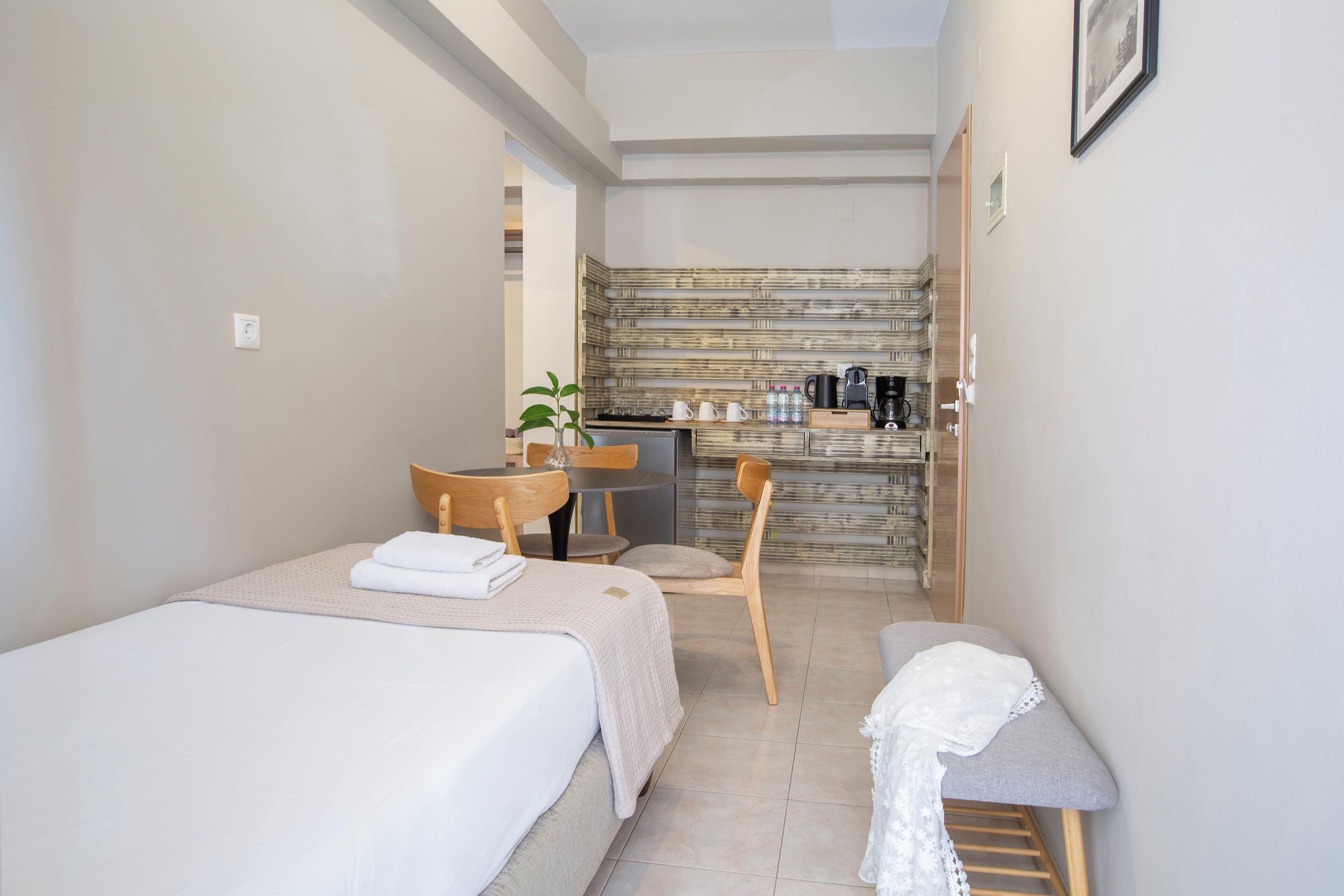 A compact hotel room with two single beds, a small table with a chair, a kitchenette, and a bathroom.