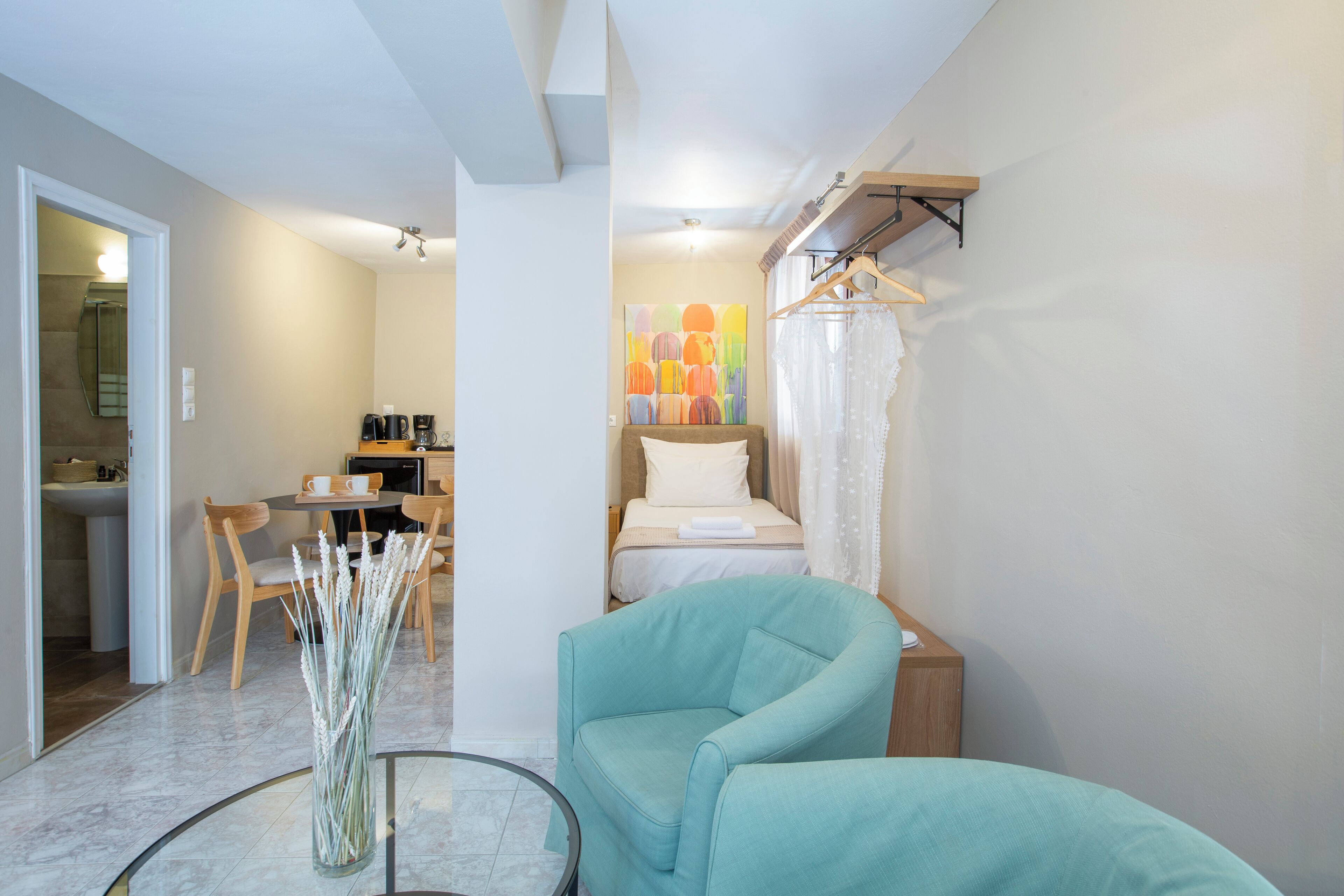 Een moderne hotelkamer met een bed, een zithoek met een stoel en een tafel, een kleine kitchenette, en een badkamer die door een open deur zichtbaar is.