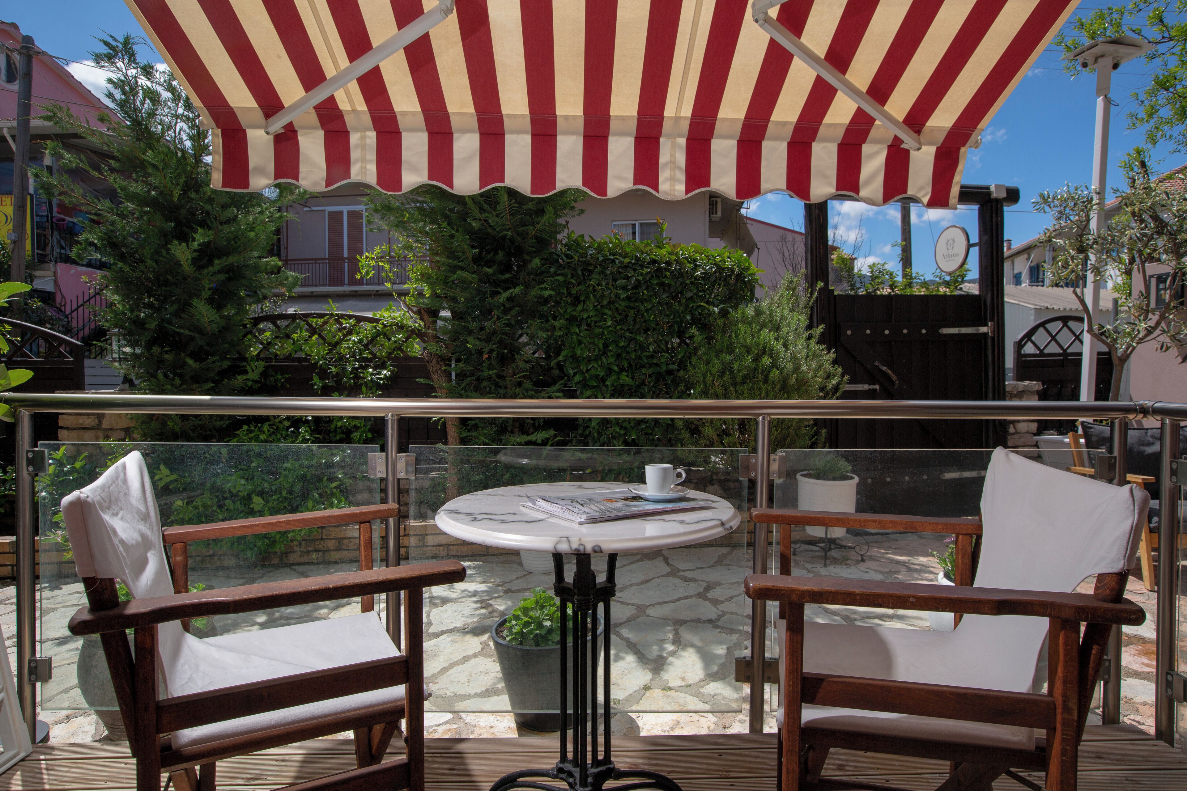 A balcony with a glass railing, two wooden chairs, a table with a white cup, and a striped awning.
