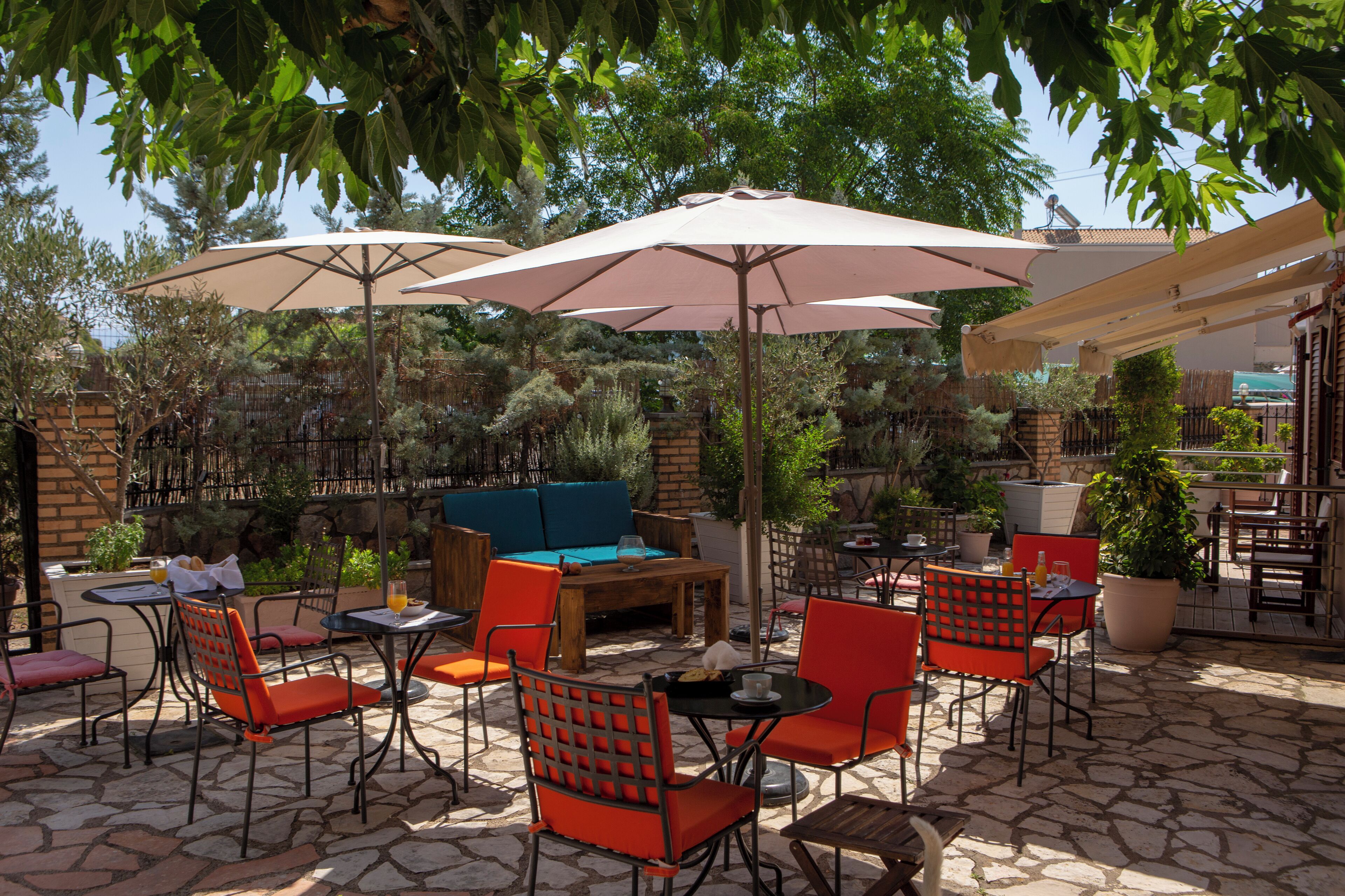 An outdoor patio with stone flooring, red and black chairs, a wooden table, and beige umbrellas providing shade.