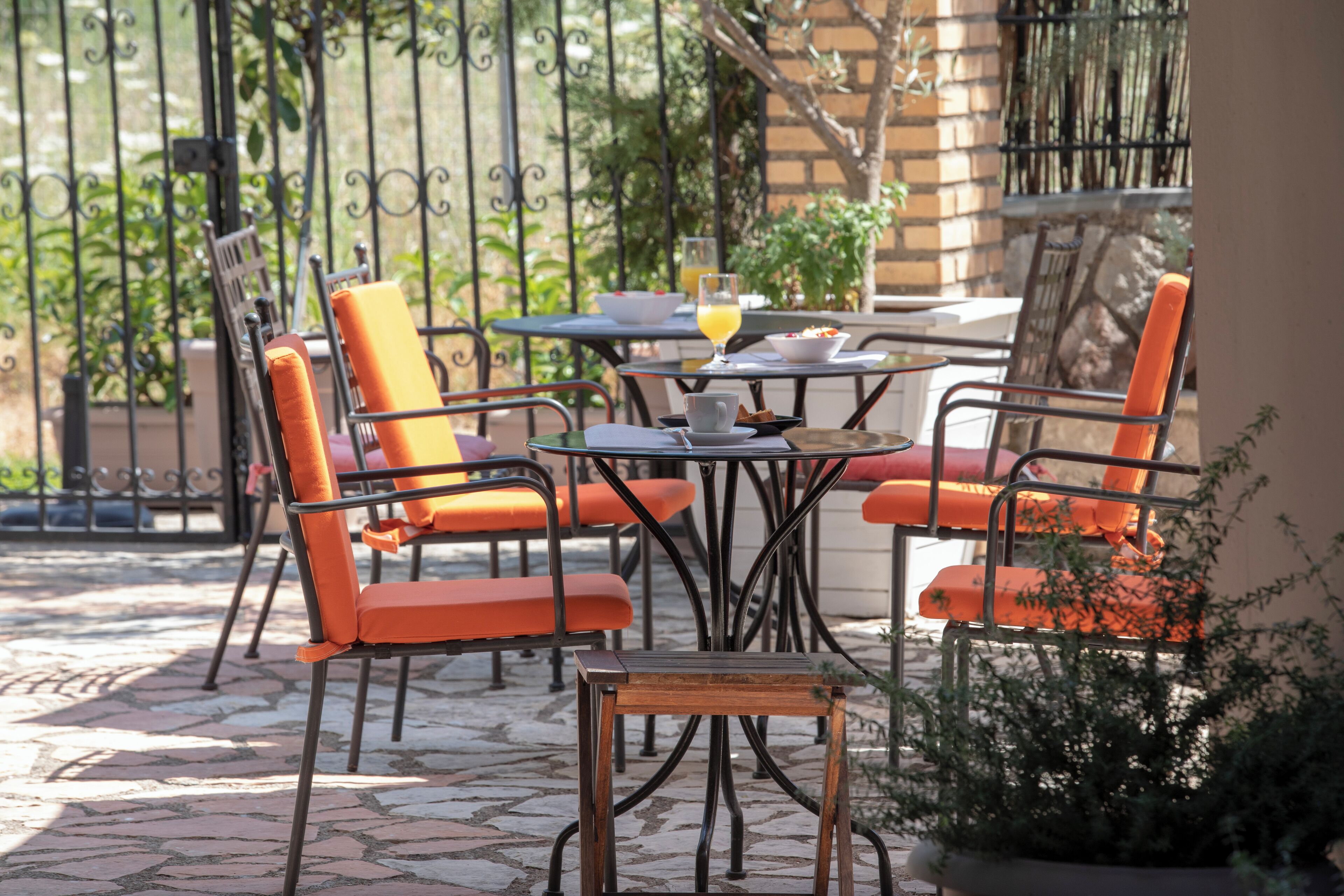 An outdoor patio with orange chairs and a small table, surrounded by a wrought iron fence and potted plants.