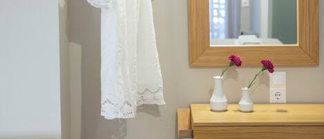 Une pièce avec un bureau en bois, une chaise, une robe blanche suspendue, un miroir et deux vases de fleurs.