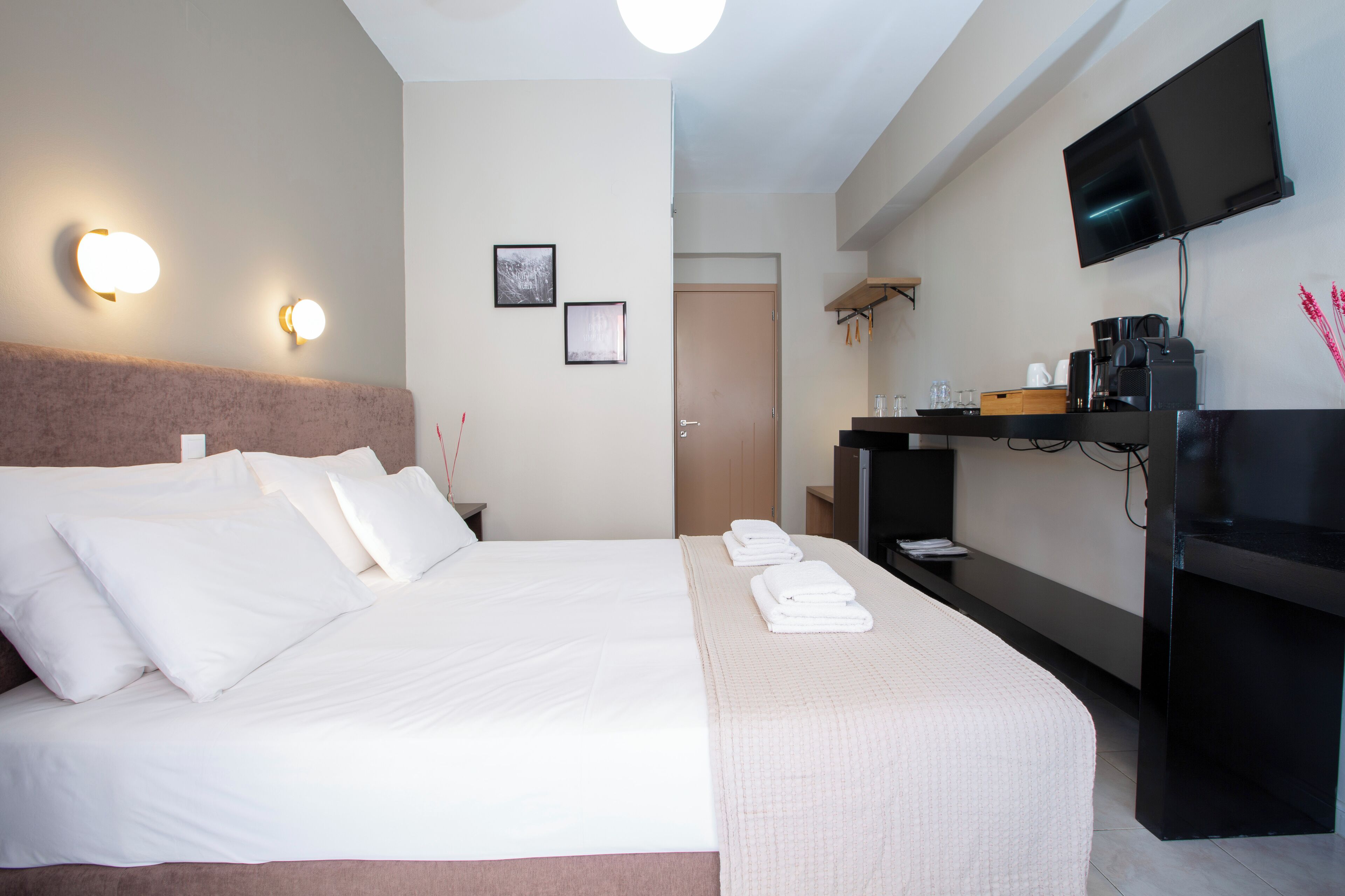 A hotel room with a large bed, a flat-screen TV mounted on the wall, a coffee machine, and a small table with a tray of folded towels.