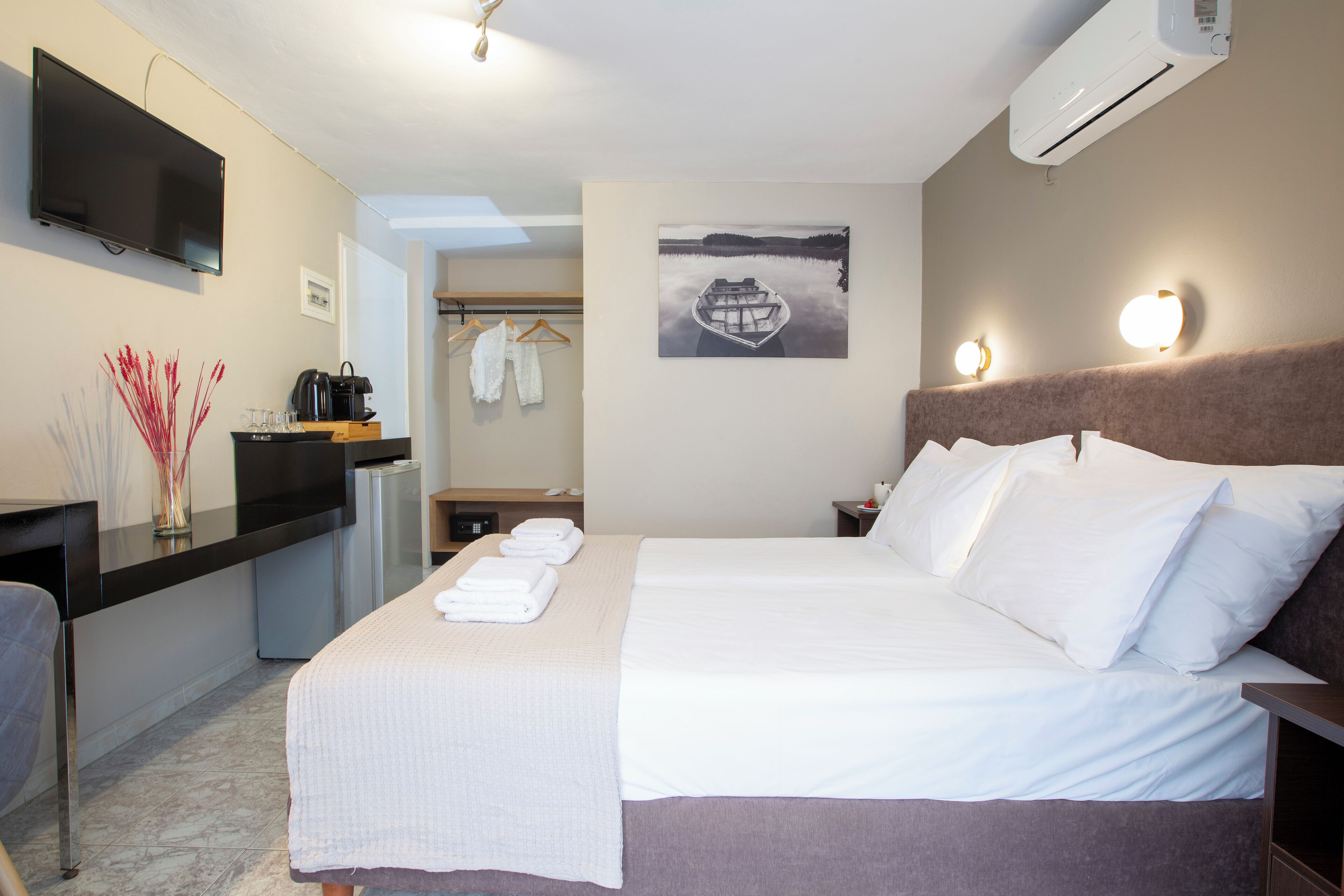 Una habitación de hotel con cama, escritorio con televisor de pantalla plana, cafetera y un cuadro de un bote en la pared.