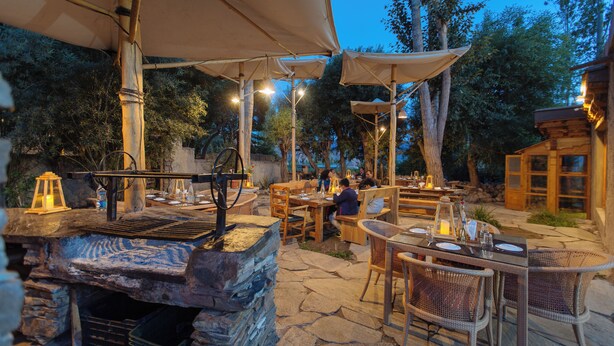 Un área de comedor al aire libre con suelo de piedra, mesas y sillas de madera, y sombrillas que dan sombra.