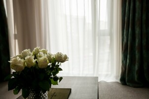 A vase with white roses on a table near a window with sheer curtains.