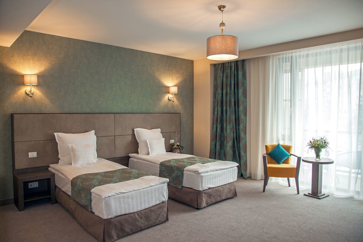 A hotel room with two beds, a chair, a small table, and a large window with curtains.