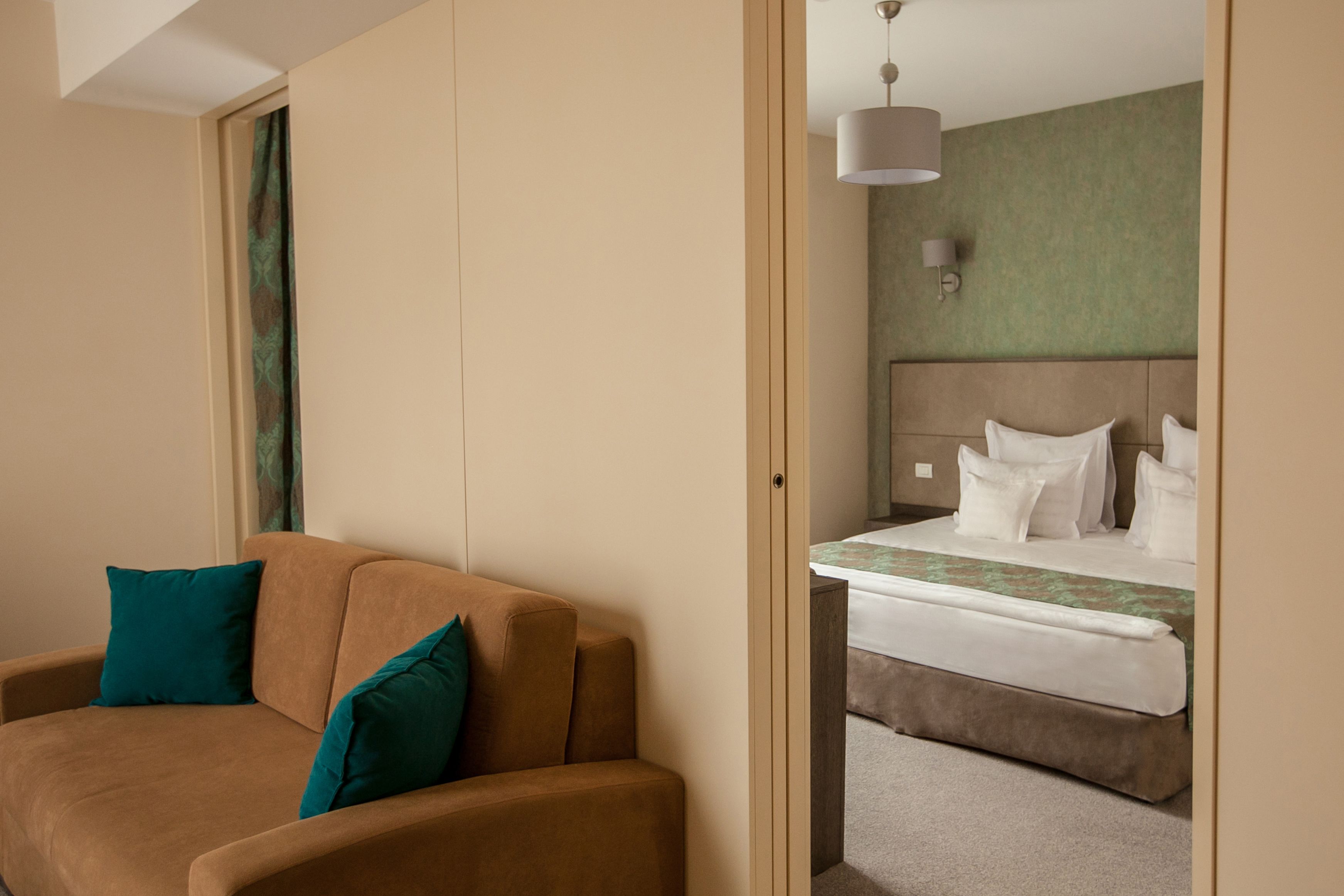 Una habitación de hotel con un sofá marrón, almohadas color turquesa, una cama con ropa de cama blanca y una pared verde.