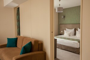 Una habitación de hotel con un sofá marrón, almohadas color turquesa, una cama con ropa de cama blanca y una pared verde.