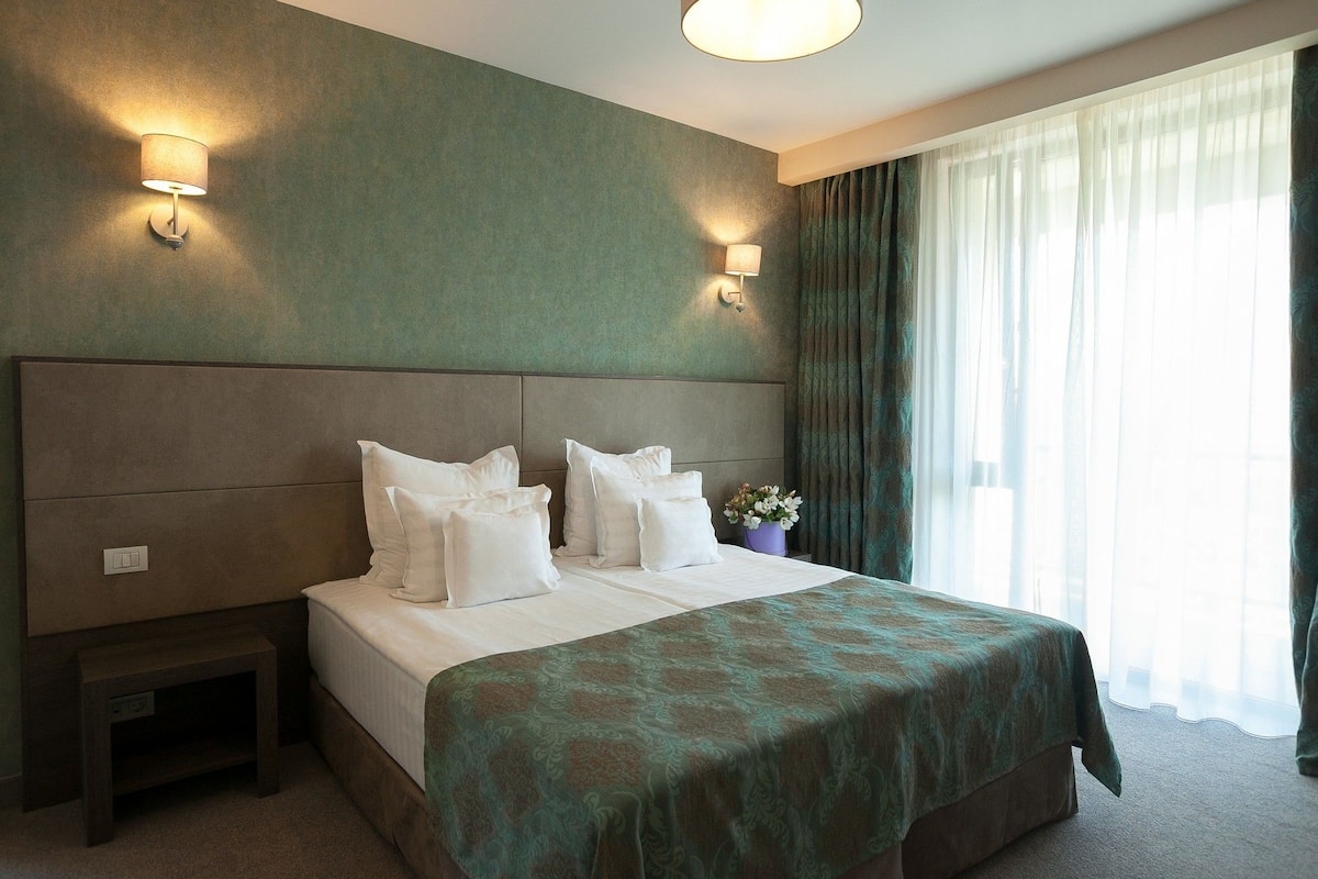 A hotel room with a large bed, two bedside lamps, a nightstand, and a window with curtains.