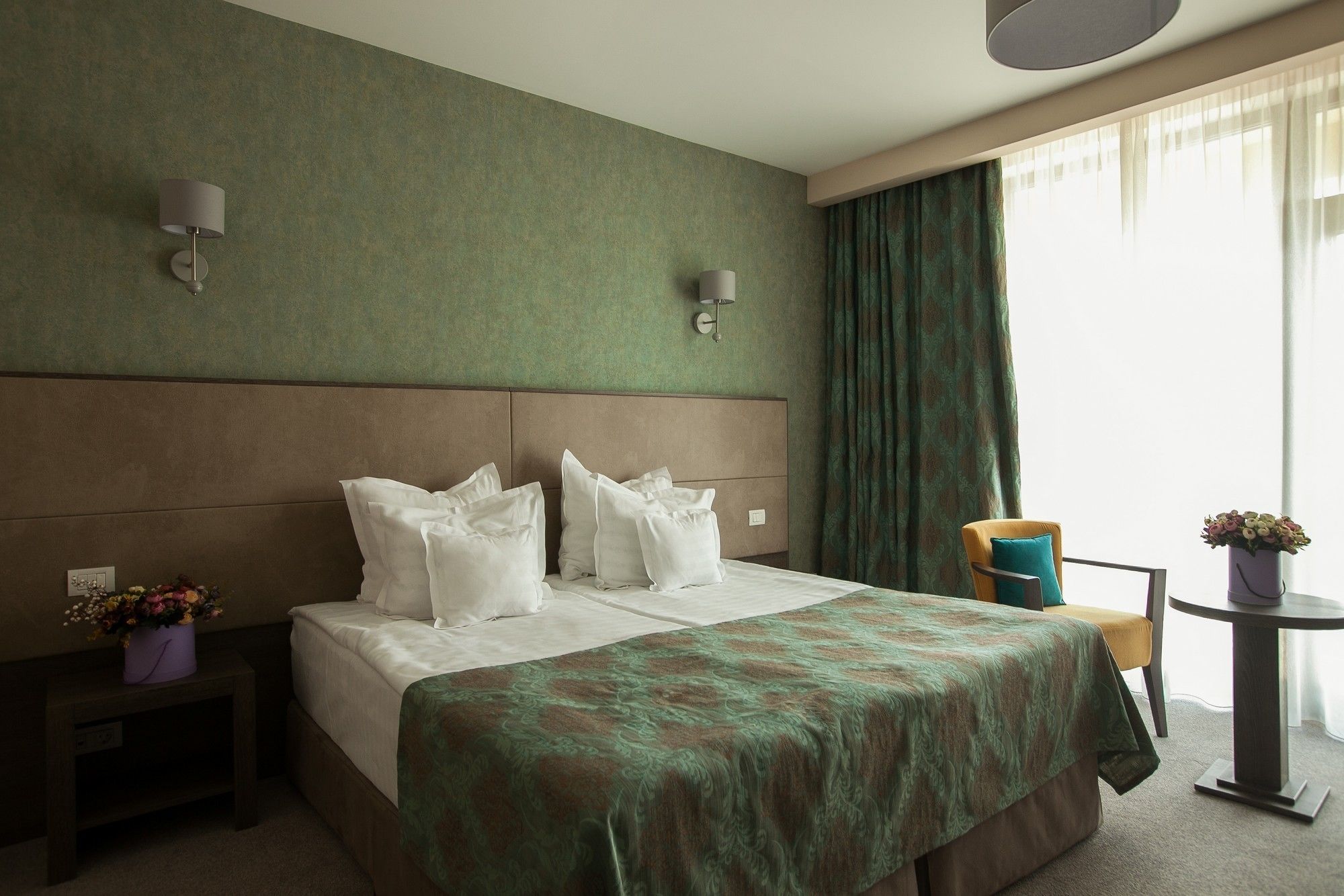 Ein Hotelzimmer mit einem großen Bett, einem Kopfteil, einem Nachttisch mit Blumenvase, einem Sessel und einem kleinen Tisch ebenfalls mit Blumenvase.