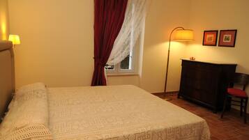 A neatly made bed with a patterned bedspread, a standing floor lamp, a wooden dresser, and two framed pictures on the wall.