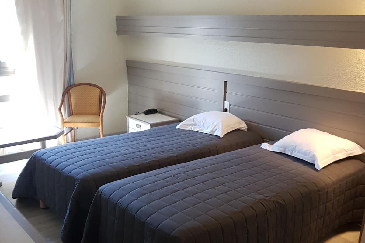 Habitación de hotel con dos camas, una mesita de noche, una silla y una ventana con cortinas.