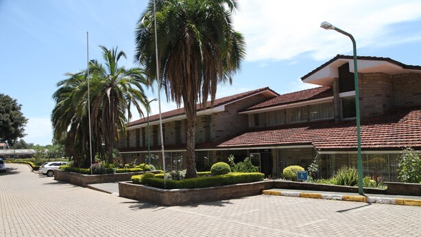 A building with a red-tiled roof, stone facade, and a paved driveway with palm trees and landscaped areas. - ARC Egerton Hotel (Njoro)