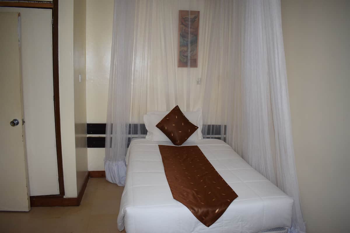 Una cama bien tendida con una colcha marrón y almohadas, una cortina blanca y una puerta a la izquierda.