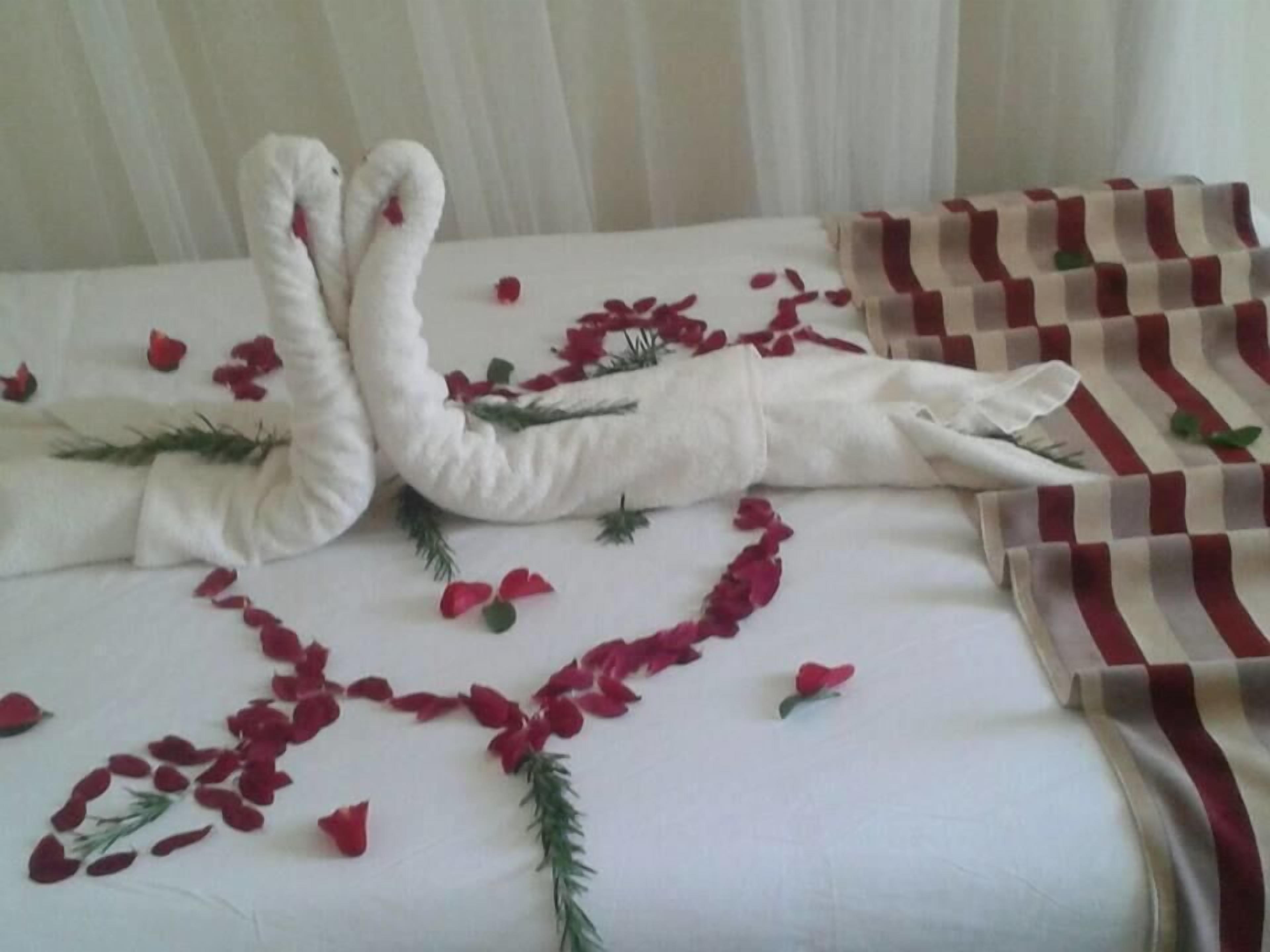 A bed with a white bedspread, red rose petals, and a white towel arranged in a swan shape.