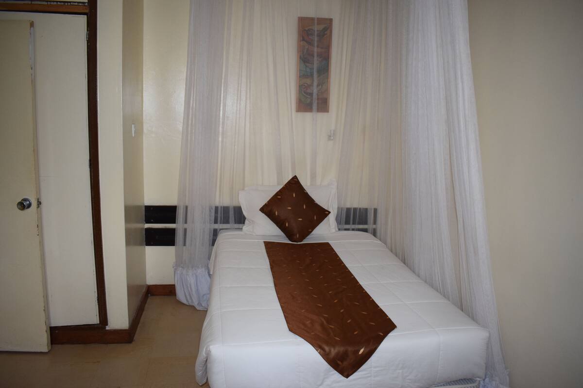 Una cama bien hecha con un cojín decorativo marrón y una colcha blanca, una puerta a la izquierda y cortinas translúcidas cubriendo la ventana.