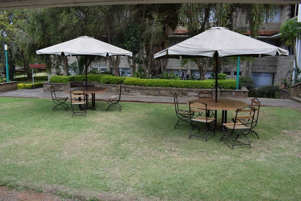 An outdoor seating area with tables and chairs under umbrellas, surrounded by greenery and a building in the background.