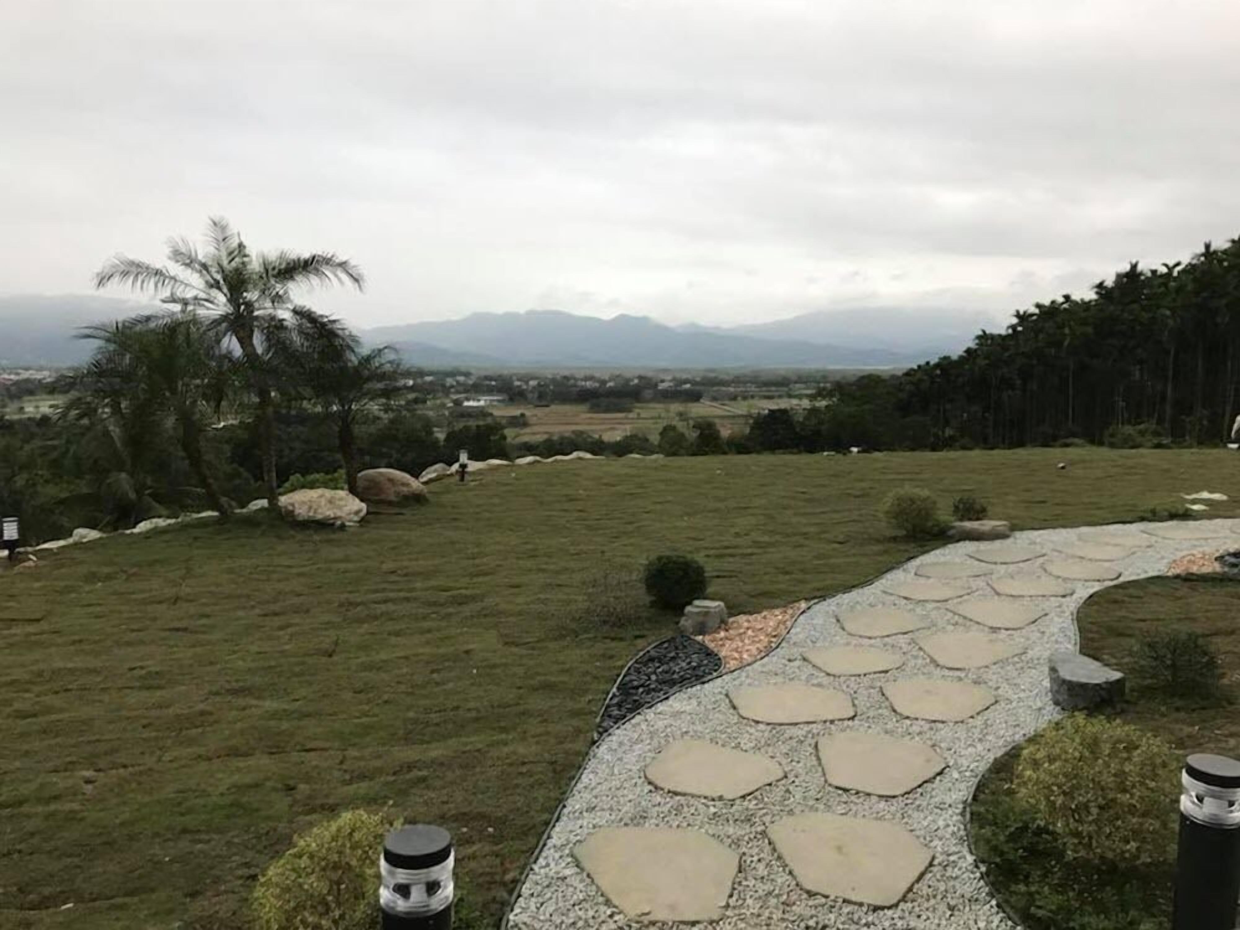 Un sendero de piedra serpentea por un área cubierta de césped, con una palmera y rocas, con vistas a un paisaje lejano con montañas.