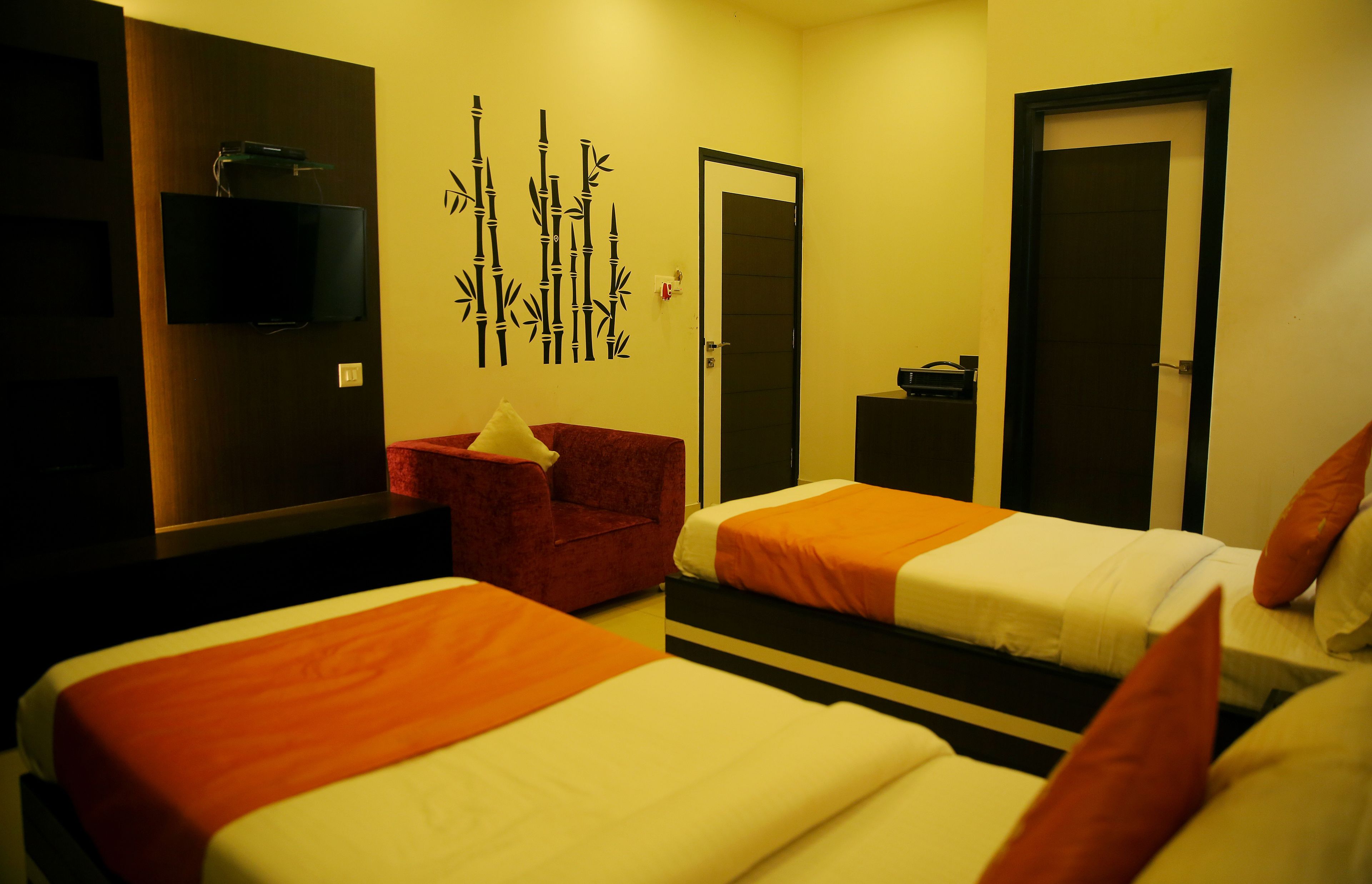A hotel room with two beds, a TV mounted on the wall, a red chair, and a door with a small table beside it.