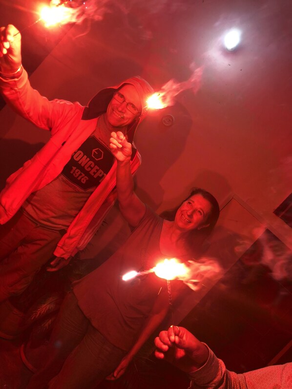 Dos personas sosteniendo bengalas, una usando una chaqueta con logotipo y texto, en un ambiente interior tenuemente iluminado.