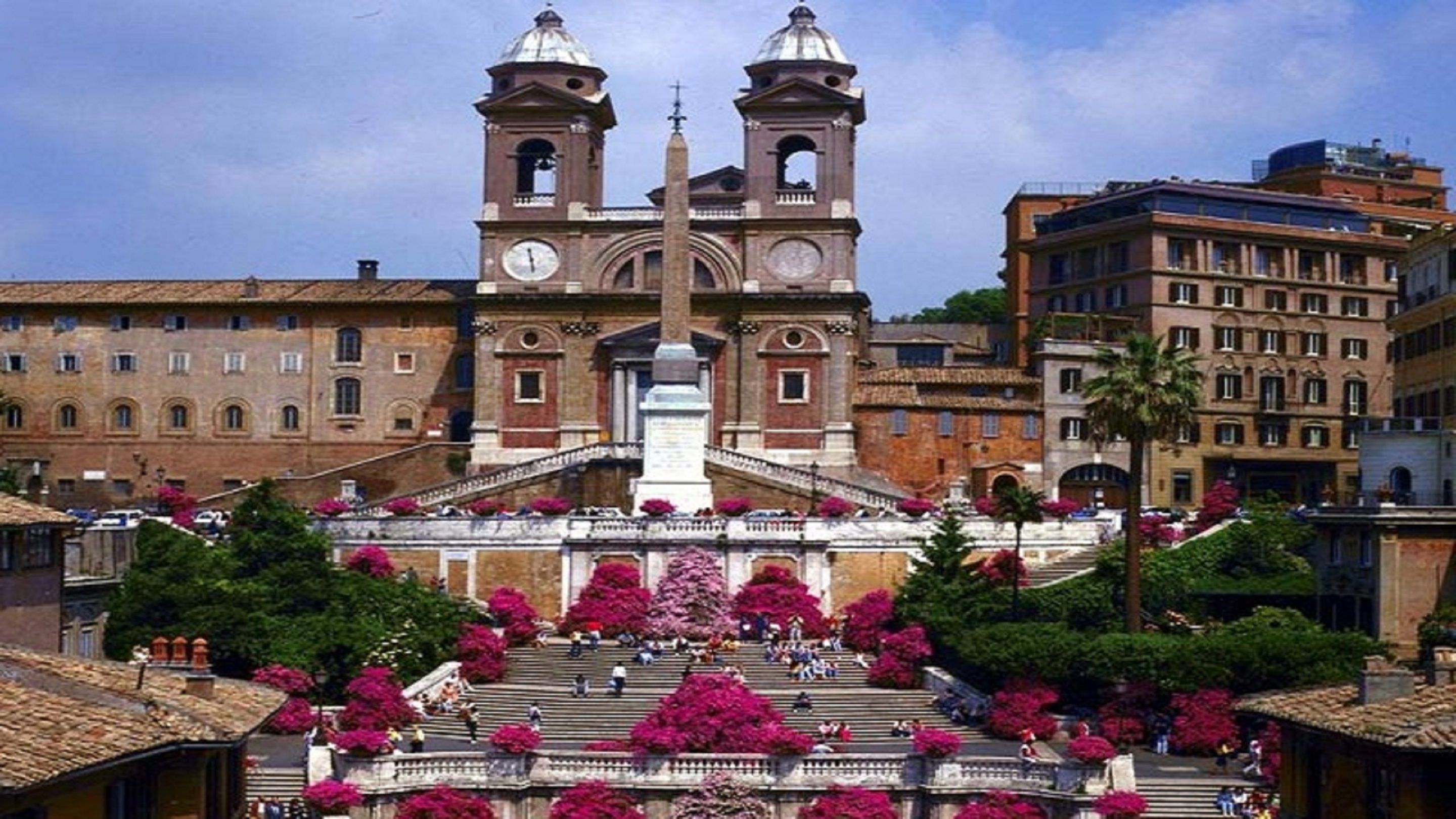 A historic church with two bell towers and a central dome, surrounded by a staircase with pink flowering bushes.