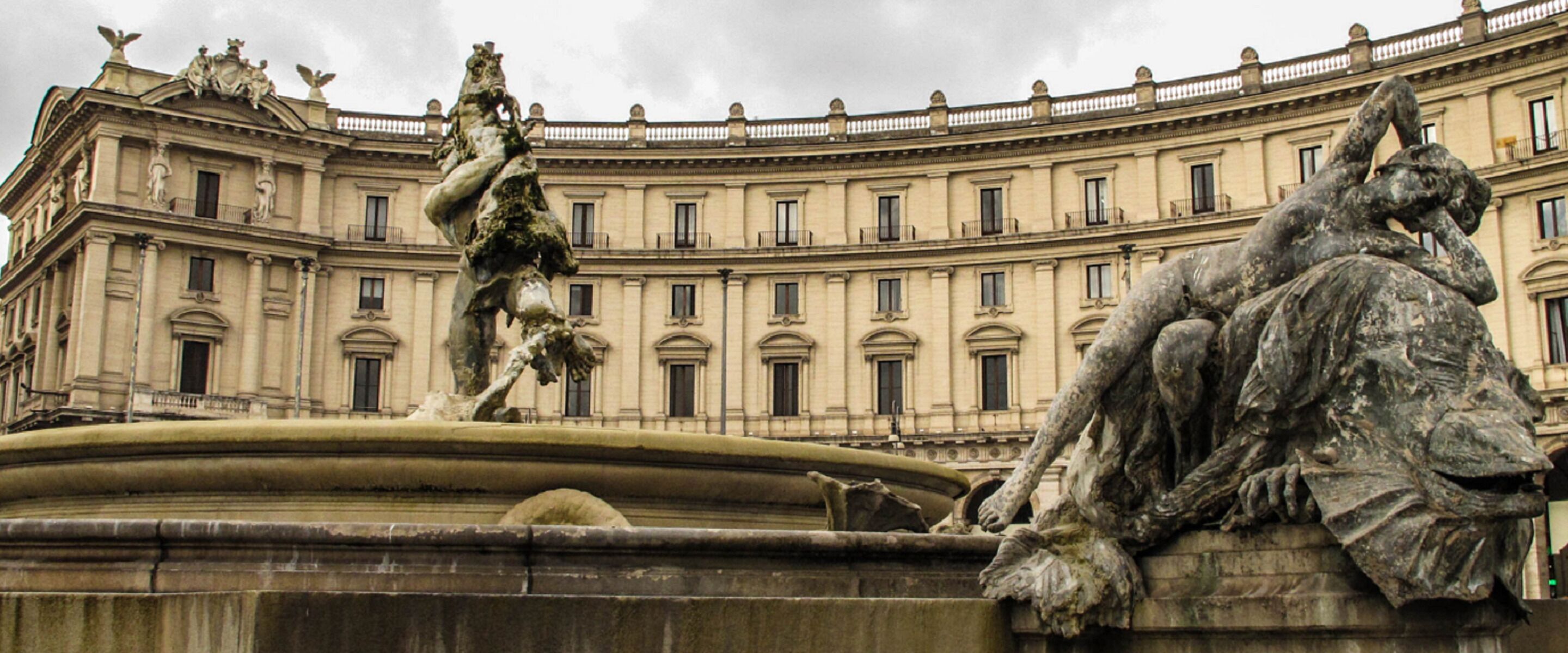Ein klassischer Brunnen mit Statuen vor einem großen, verzierten Gebäude mit zahlreichen Fenstern und Balkonen.