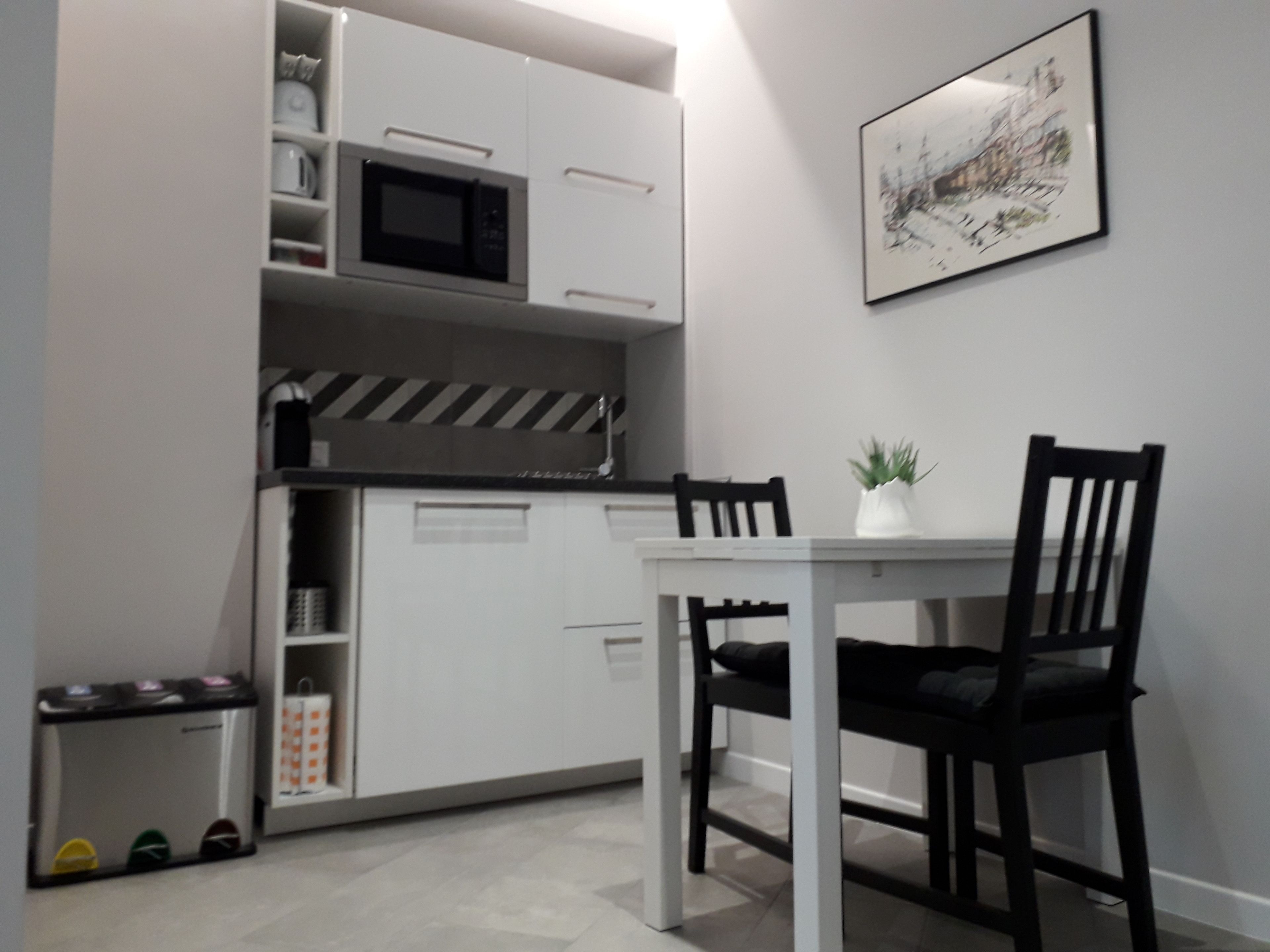 Een kleine keuken met een magnetron, een kleine koelkast en een prullenbak. Er staat een eettafel met twee stoelen en op de muur staat een plant in pot.