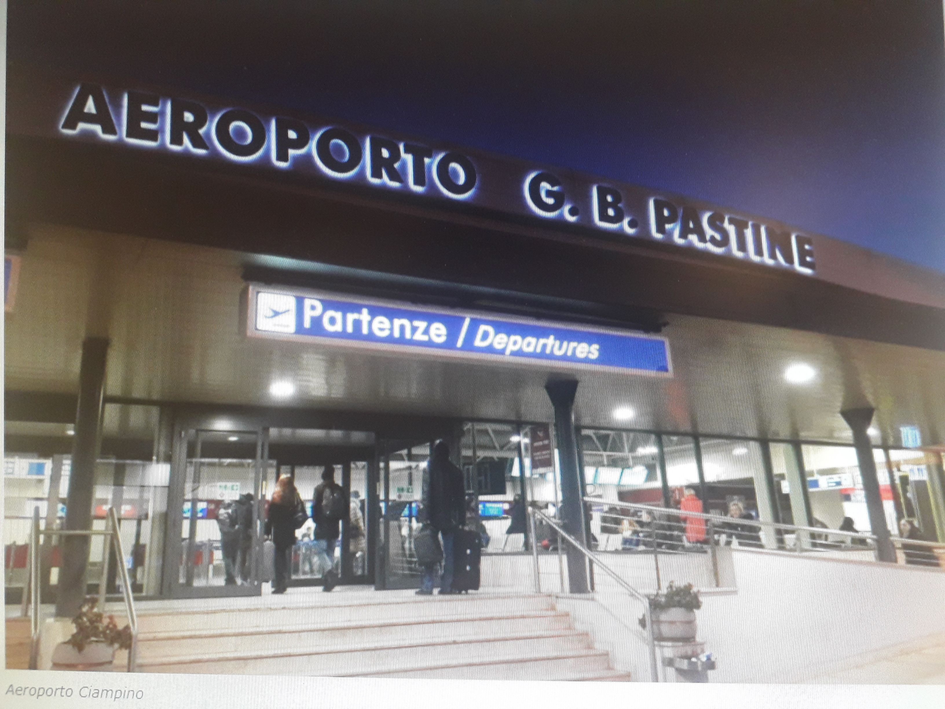Una terminal de aeropuerto con señalización que indica 'AEROPORTO G.B. PASTINE' y 'Partenze / Departures'.
