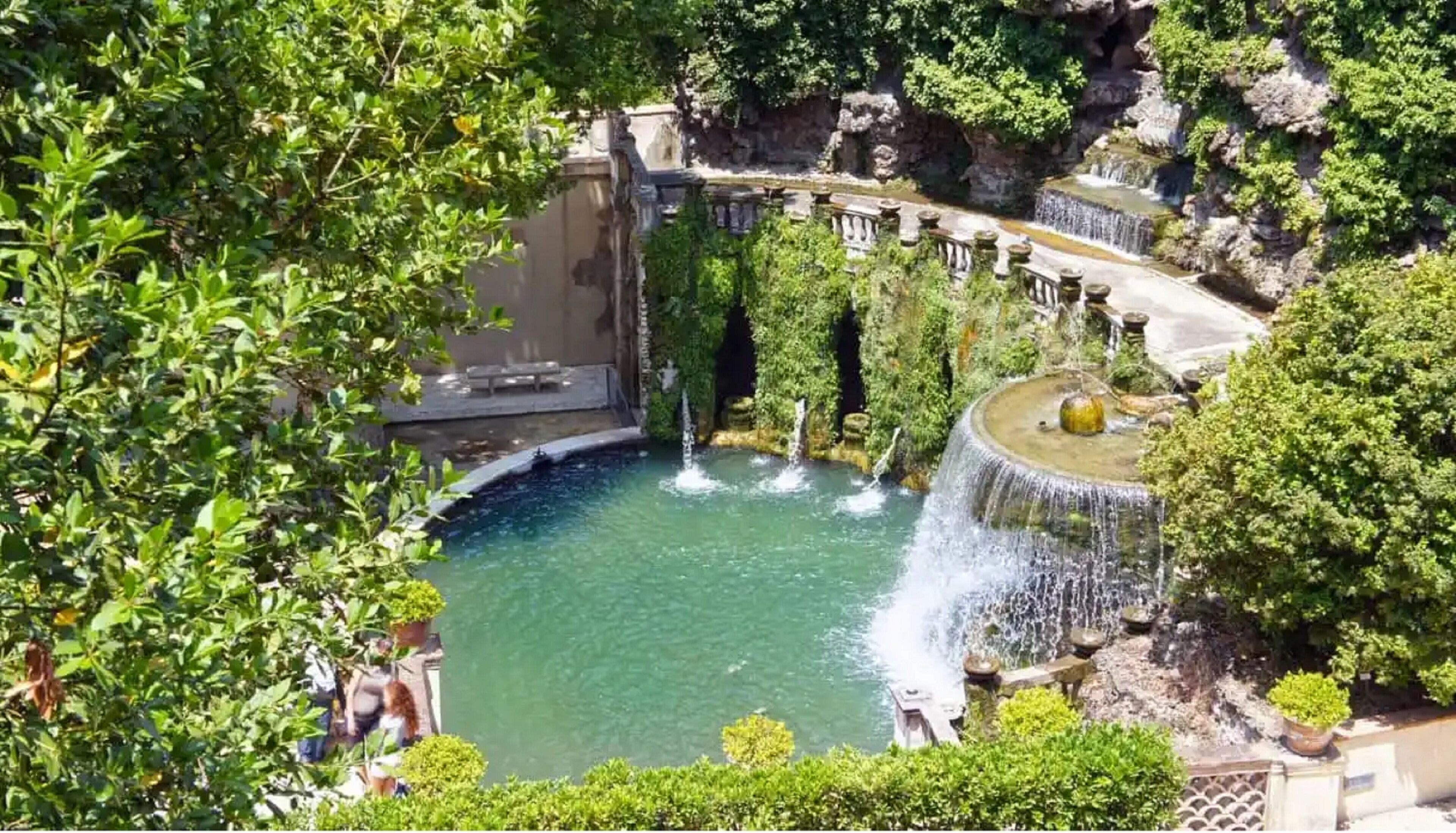 Un jardin luxuriant doté d’un élément aquatique central, comprenant un étang et des cascades, le tout entouré d’une végétation dense et de structures en pierre.