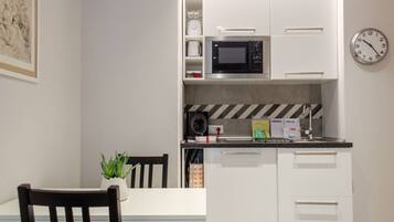 Cocina compacta con microondas, hervidor y cafetera sobre la encimera. Hay un reloj en la pared y una pequeña planta en maceta sobre la mesa.