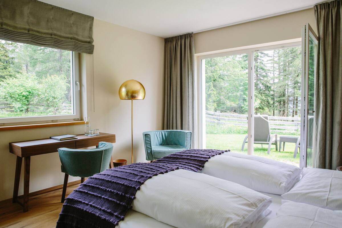 A bedroom with a bed, a desk, a chair, and a view of a garden through large windows.
