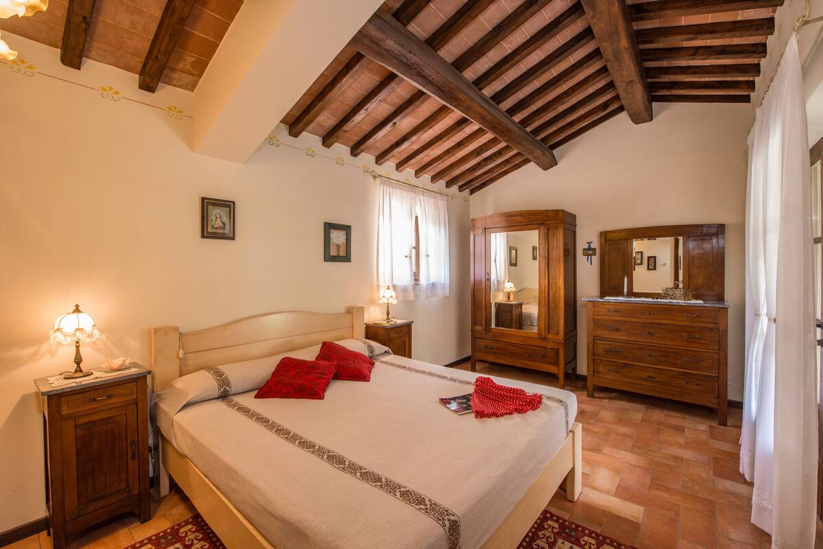 Een slaapkamer met een houten plafond, een bed met rode kussens, een nachtkastje met een lamp, een dressoir en een spiegel.