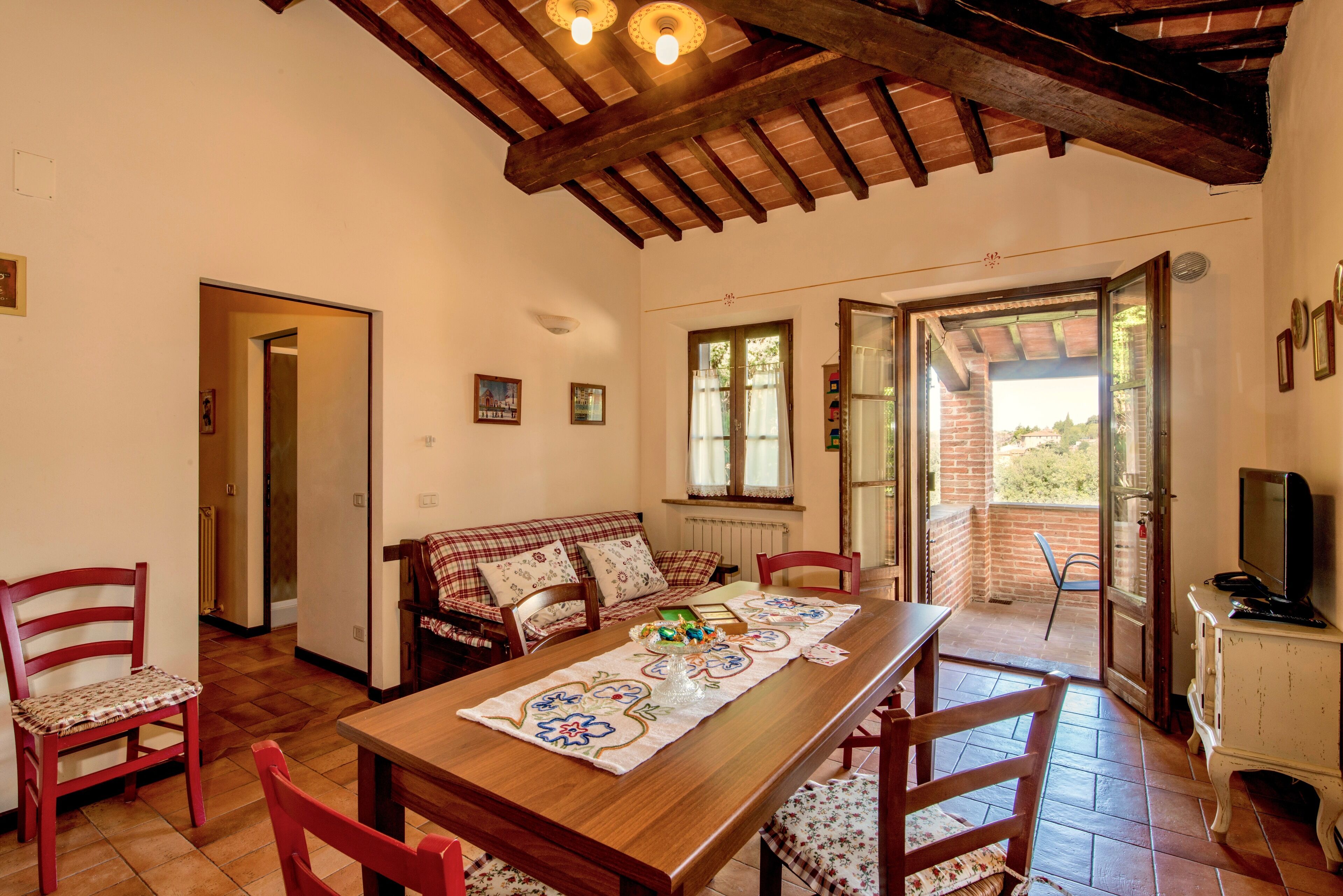 Un'accogliente sala da pranzo con un tavolo in legno, sedie rosse e una tovaglia a quadri. C'è anche un divano con cuscini decorati e un televisore su un mobile.