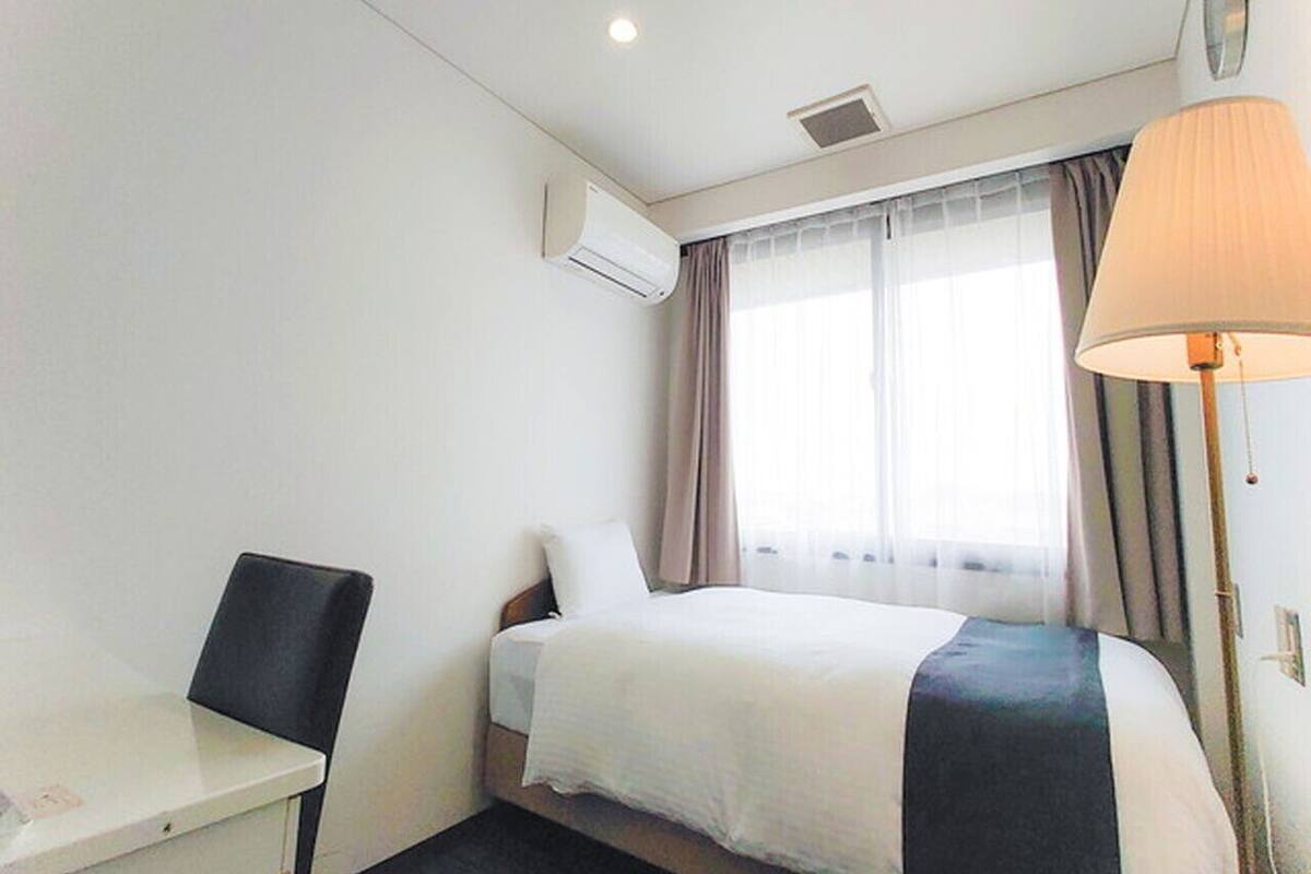 A hotel room with a bed, a desk, a chair, an air conditioning unit, a lamp, and a window with curtains.
