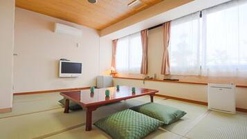 Una habitación tradicional japonesa con suelo de tatami, una mesa de madera baja, cojines, un televisor y una ventana con cortinas.