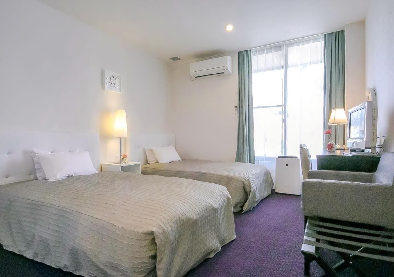 Una habitación de hotel con dos camas, un escritorio con una computadora, una silla y un ventanal con cortinas.